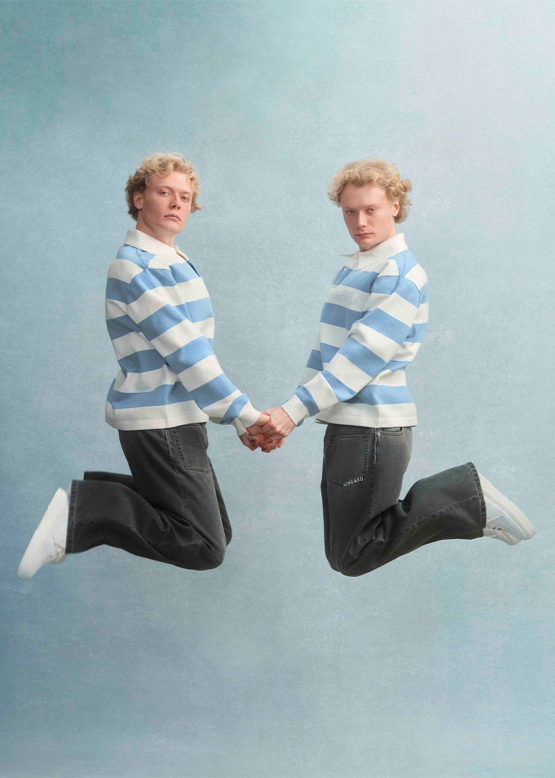 Two individuals wearing blue and white striped sweaters and jeans jump in mid-air, holding hands against a light blue background.