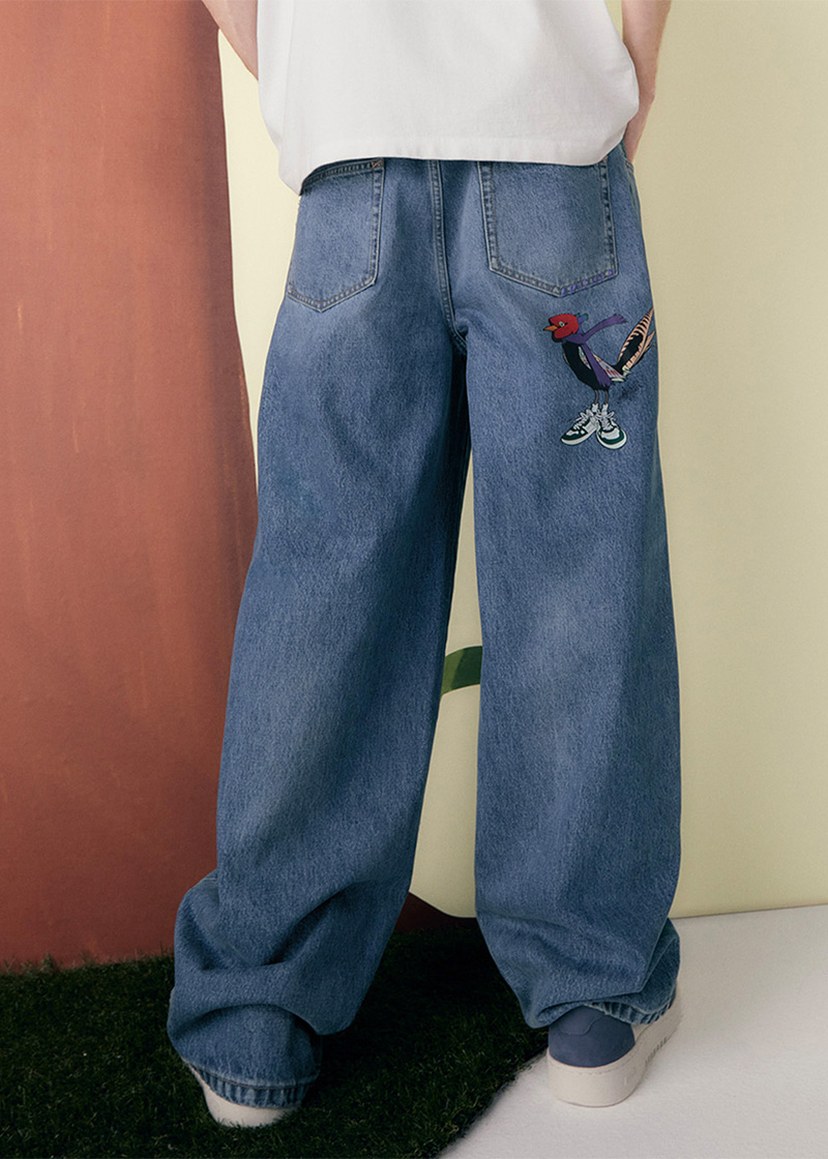 Person wearing wide-legged jeans with embroidered bird design on the back pocket, standing on a grass-like surface against a beige and brown background.