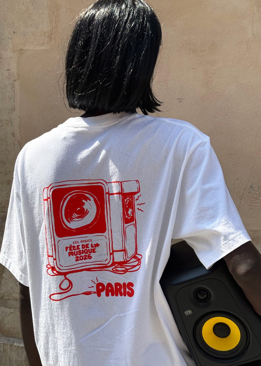 Person wearing a white T-shirt with a red speaker design and "Fête De La Musique 2026, Paris" text, holding a speaker.