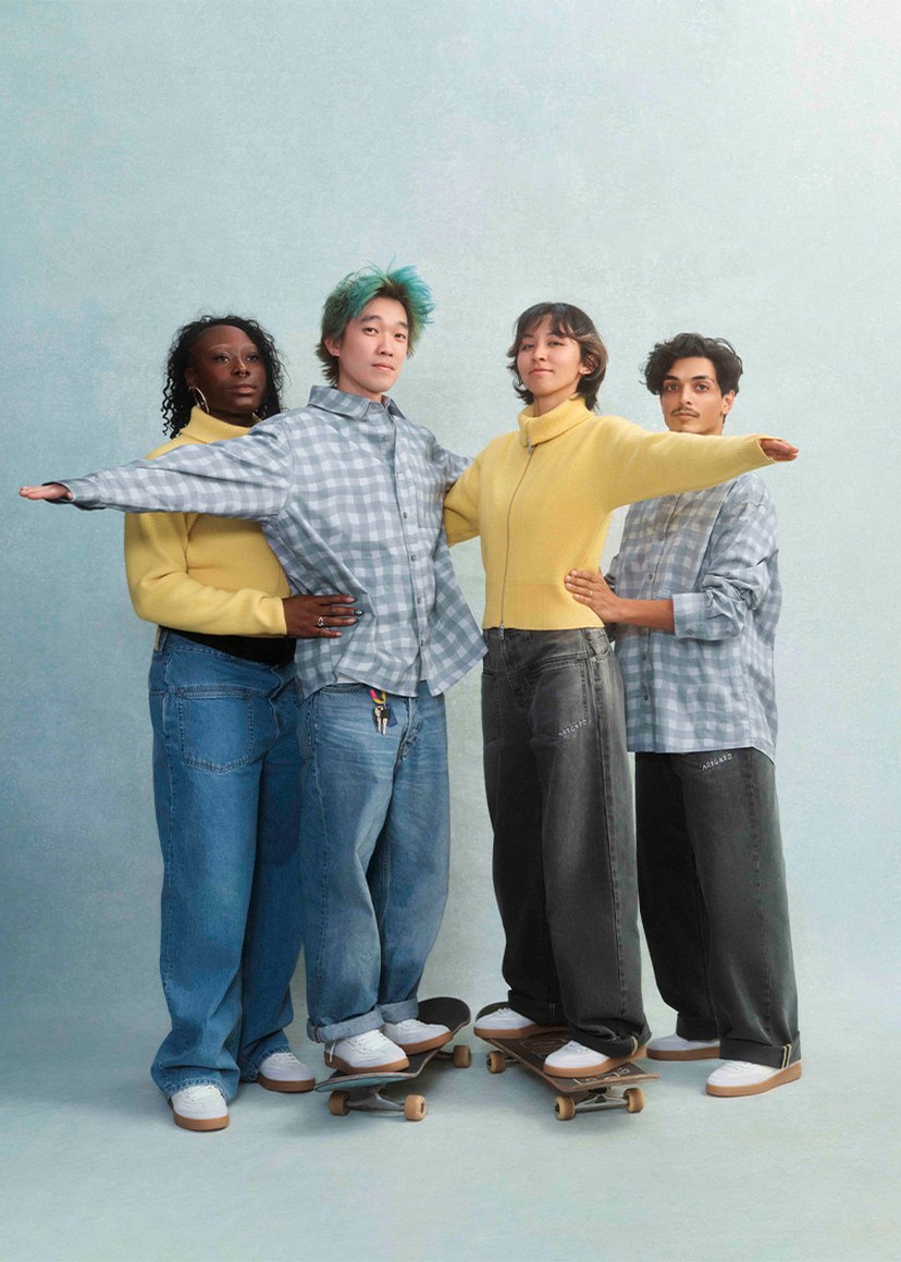 Four people posing in casual clothing, three wearing checkered shirts, two on skateboards, all standing together against a neutral backdrop.
