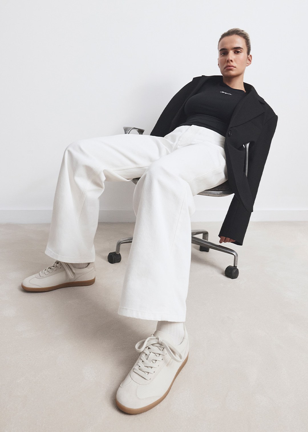 Person in a black blazer, white wide-leg pants, and beige sneakers, seated on a chair against a plain white background.