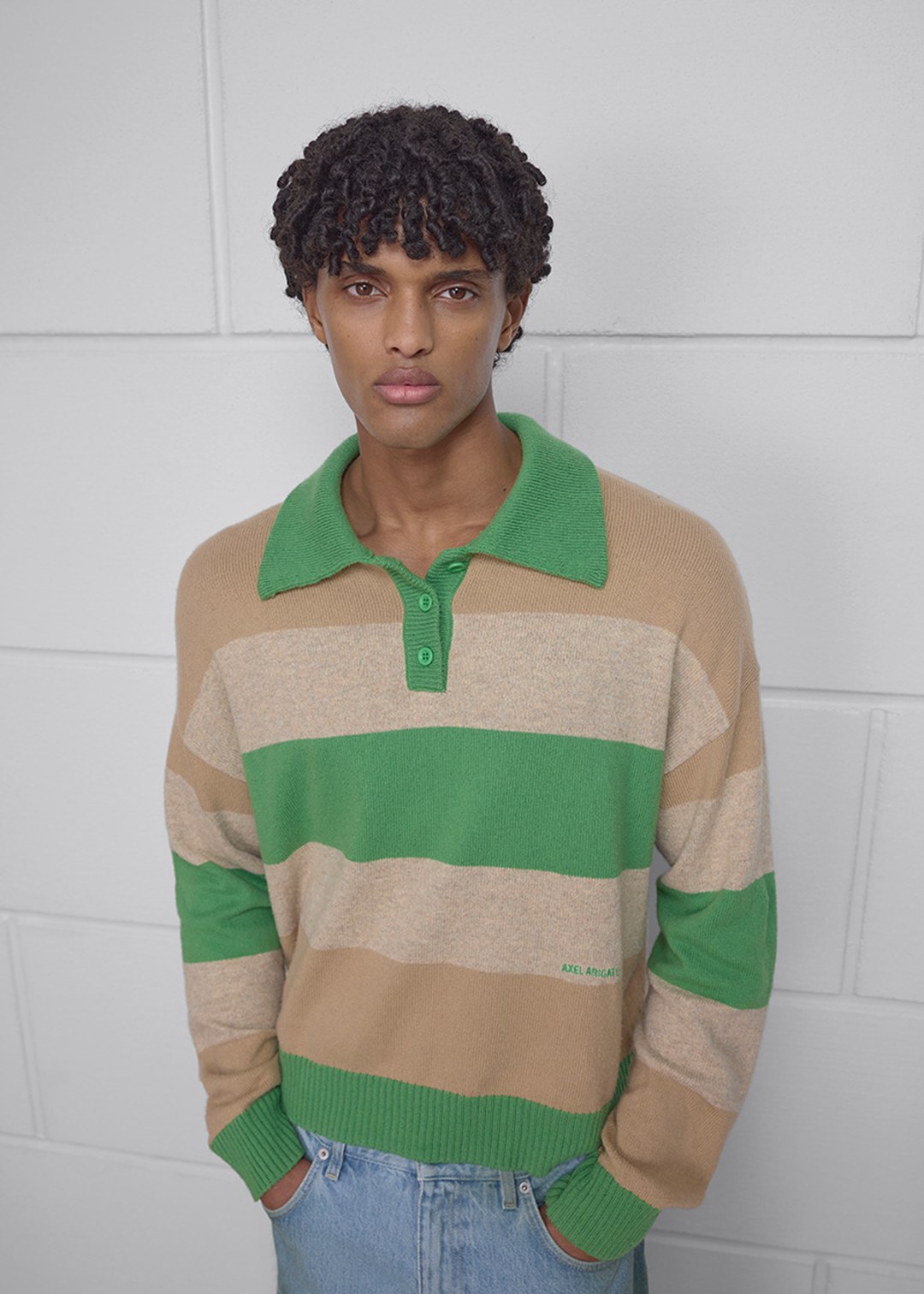 Person wearing a beige and green striped sweater with a green collar, standing against a white brick wall.