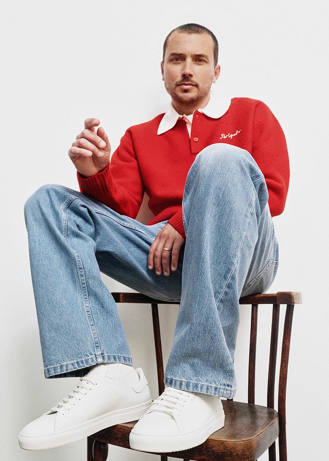 Man in a red sweater and jeans sits on a chair, wearing white sneakers, looking at the camera against a white background.