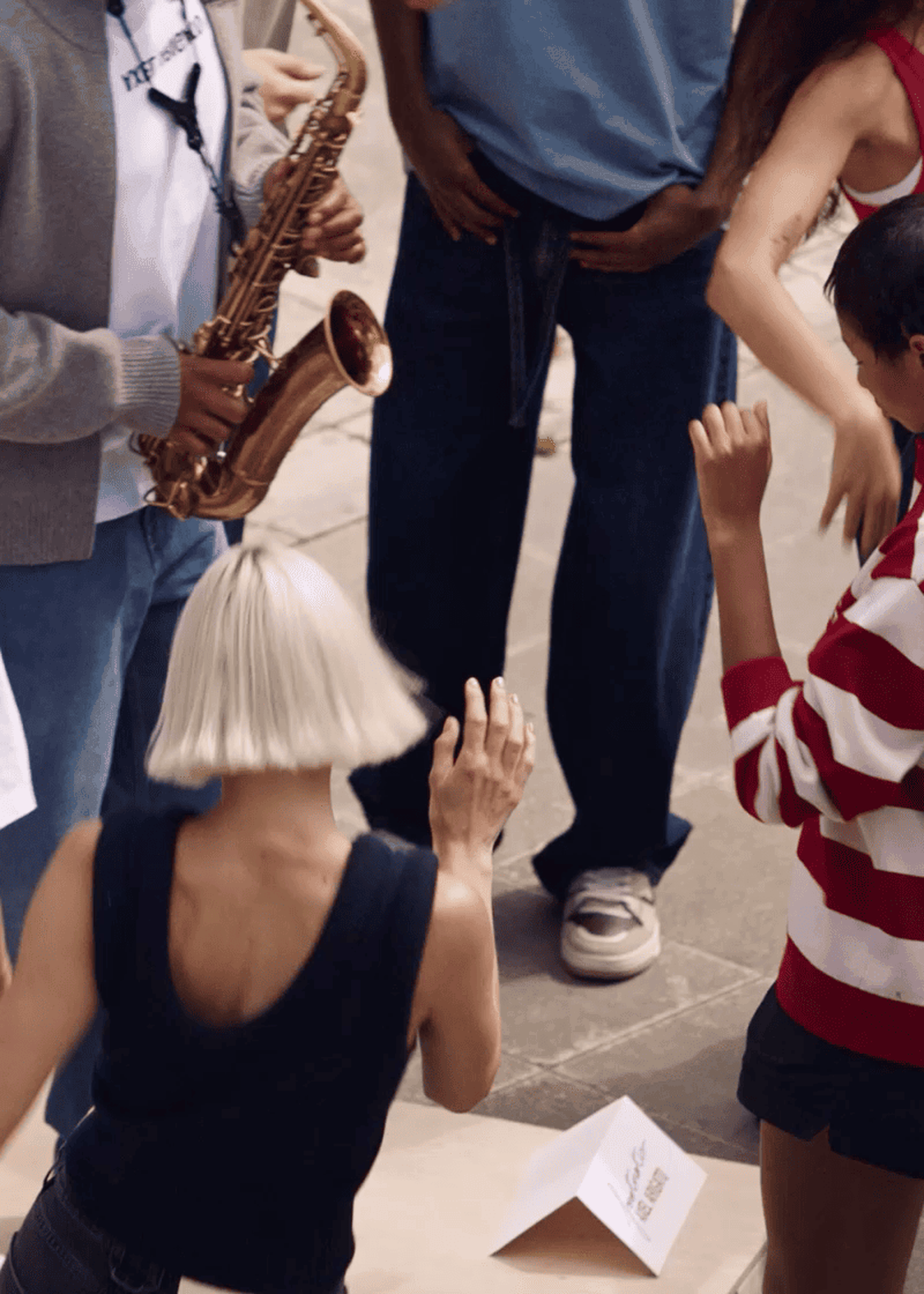 People dancing energetically outdoors, with a saxophonist playing. One person has short blonde hair, and another wears a red and white striped shirt.
