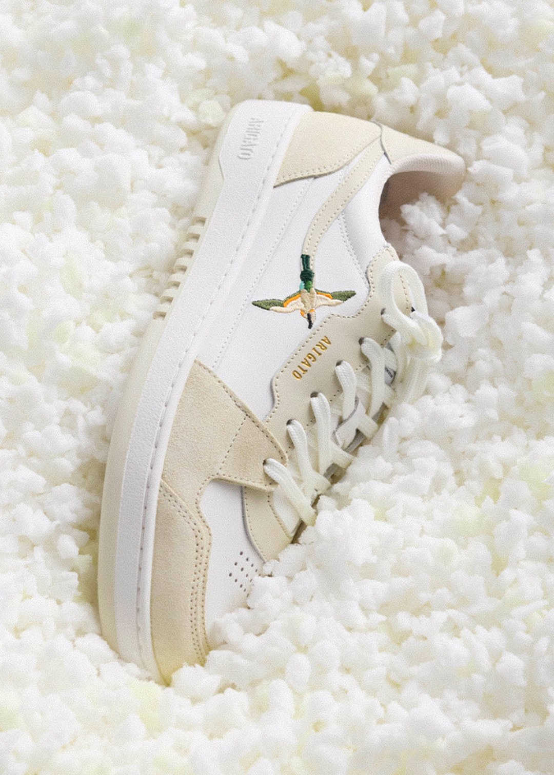 A white and beige sneaker with a green bird embroidery, set on a textured white surface.