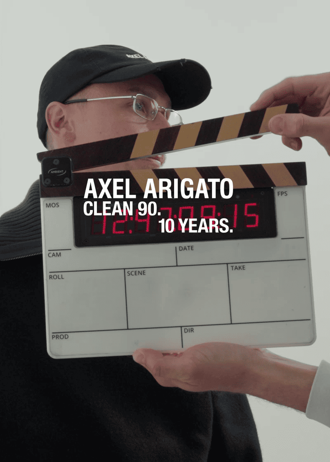 A person holding a clapperboard in front of someone wearing glasses and a cap. Text reads "Axel Arigato Clean 90. 10 Years."