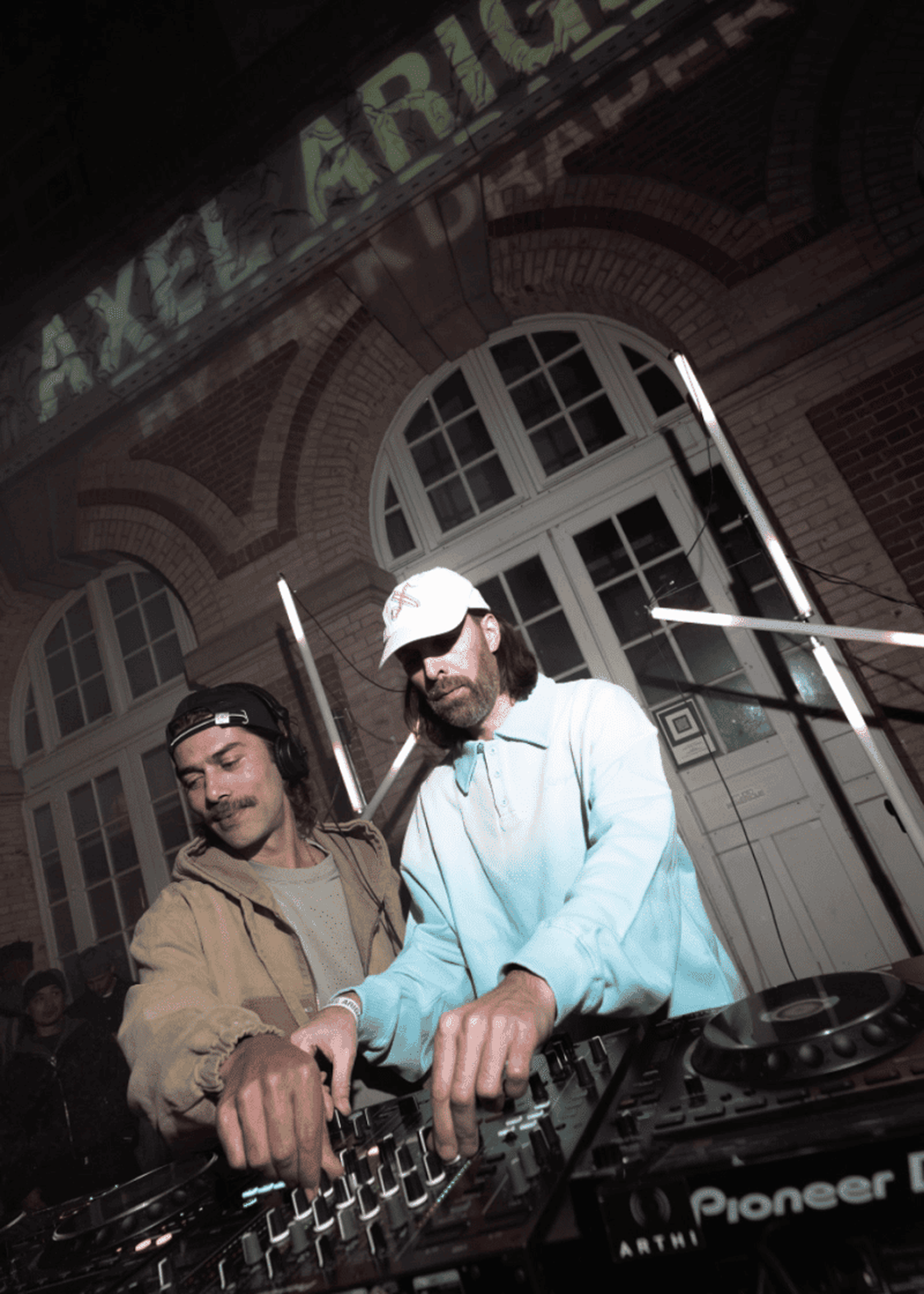 Two DJs mix music together at an event, focused on their equipment. A building with arched windows is lit in the background.