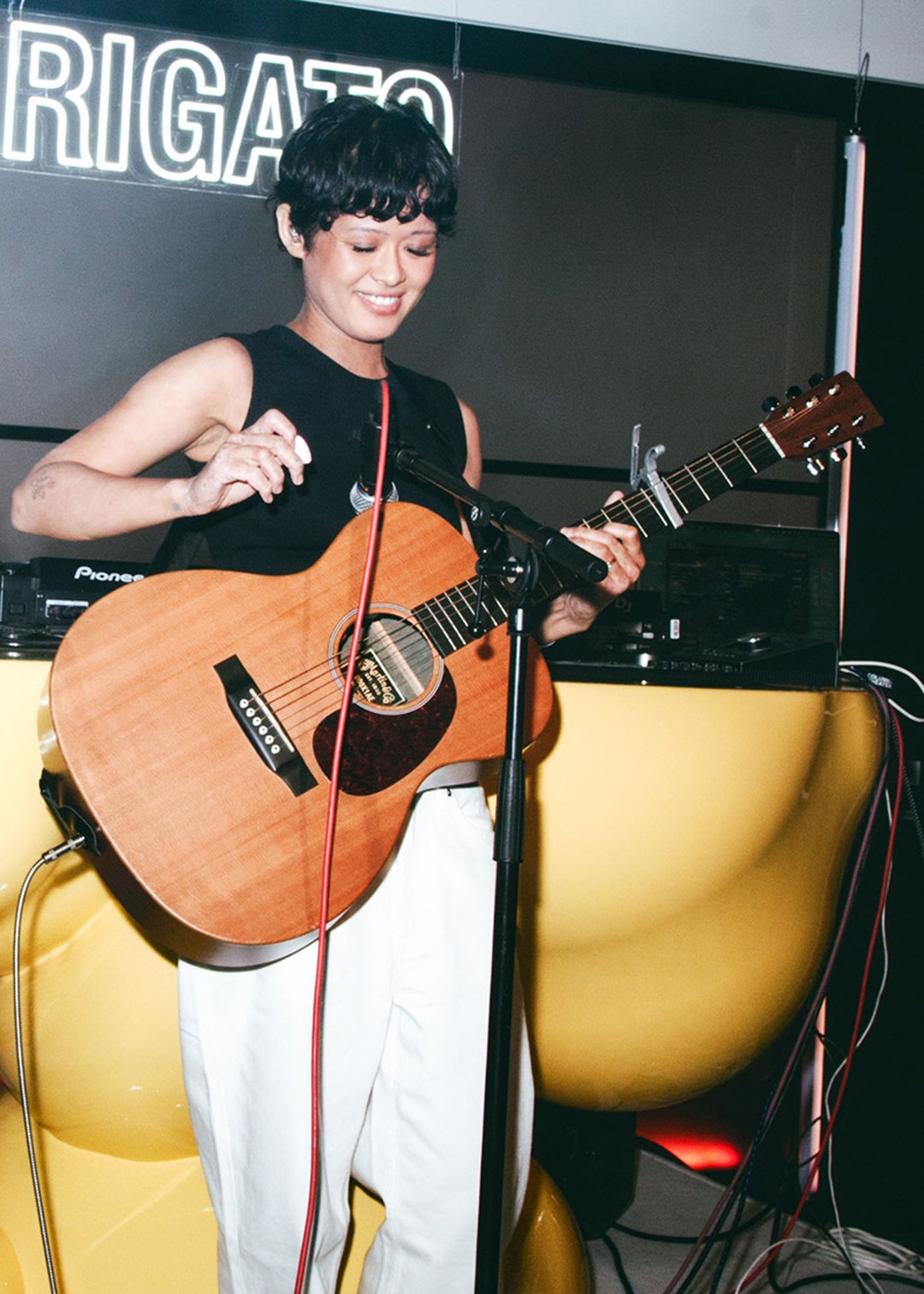 Person smiling while playing an acoustic guitar on stage, standing in front of a yellow sculpture and lit sign.