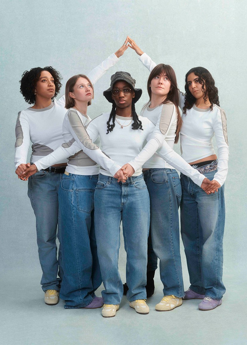 Five people stand in a circle, holding hands and wearing similar outfits with white tops and blue jeans, against a light background.