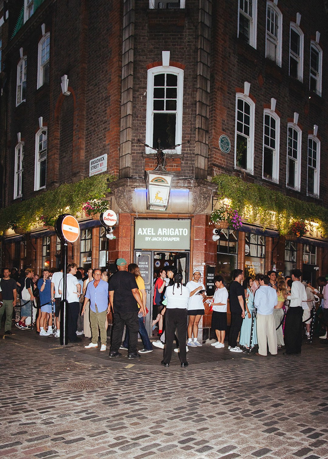 Crowd gathered outside a brick corner building with "Axel Arigato by Jack Draper" sign, adorned with hanging greenery and flowers at night.