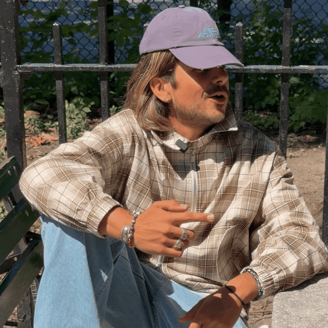 Man in plaid shirt and purple cap sits on a bench, gesturing with his hand, with a fence and leaves in the background.