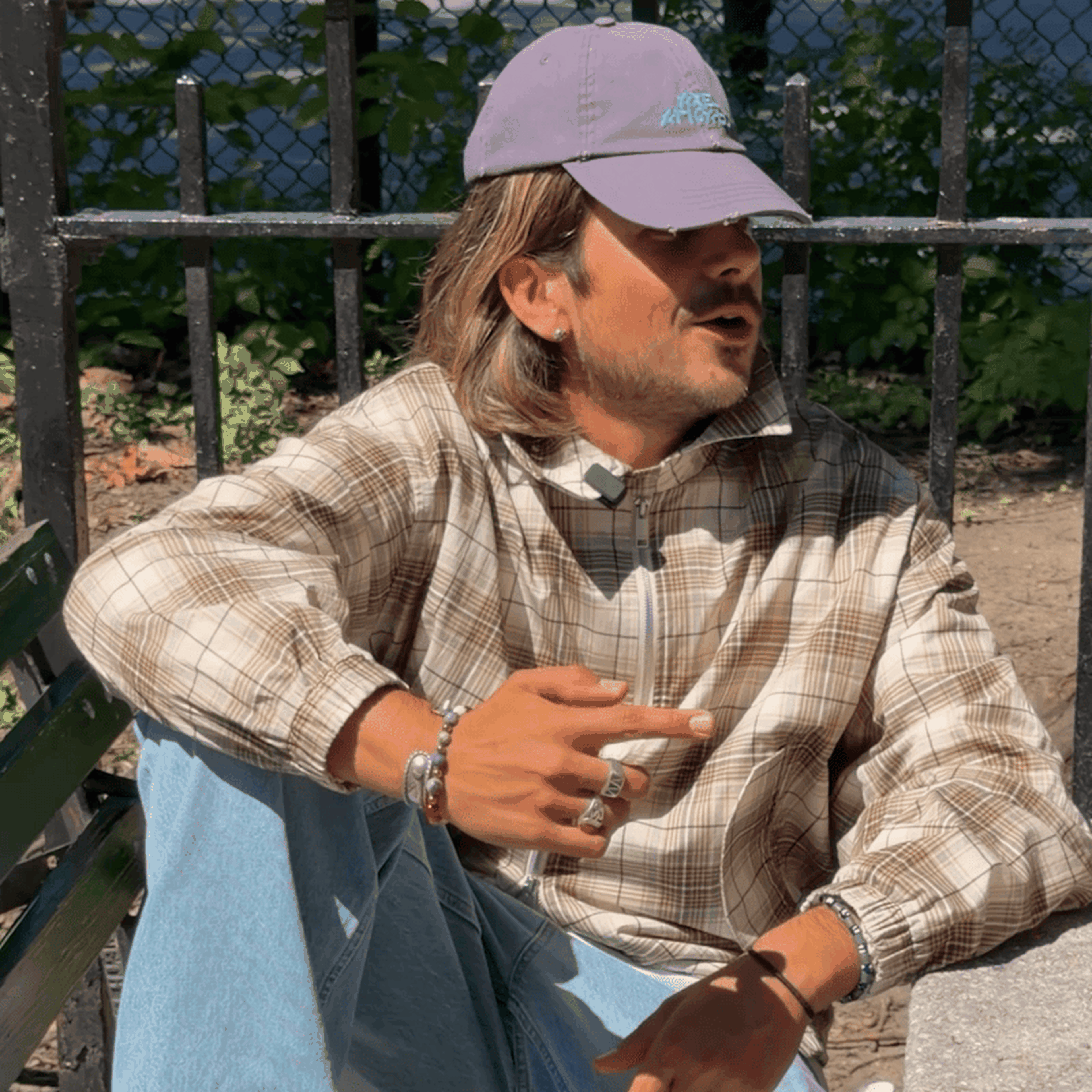 Man in plaid shirt and purple cap sits on a bench, gesturing with his hand, with a fence and leaves in the background.
