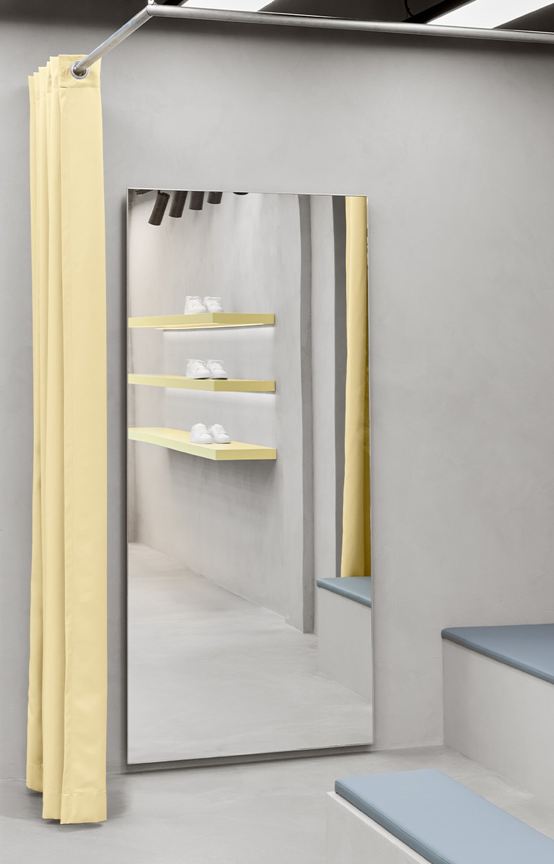 A minimalist dressing room with a large mirror, yellow curtain, gray walls, and shelves reflected, displaying white shoes.