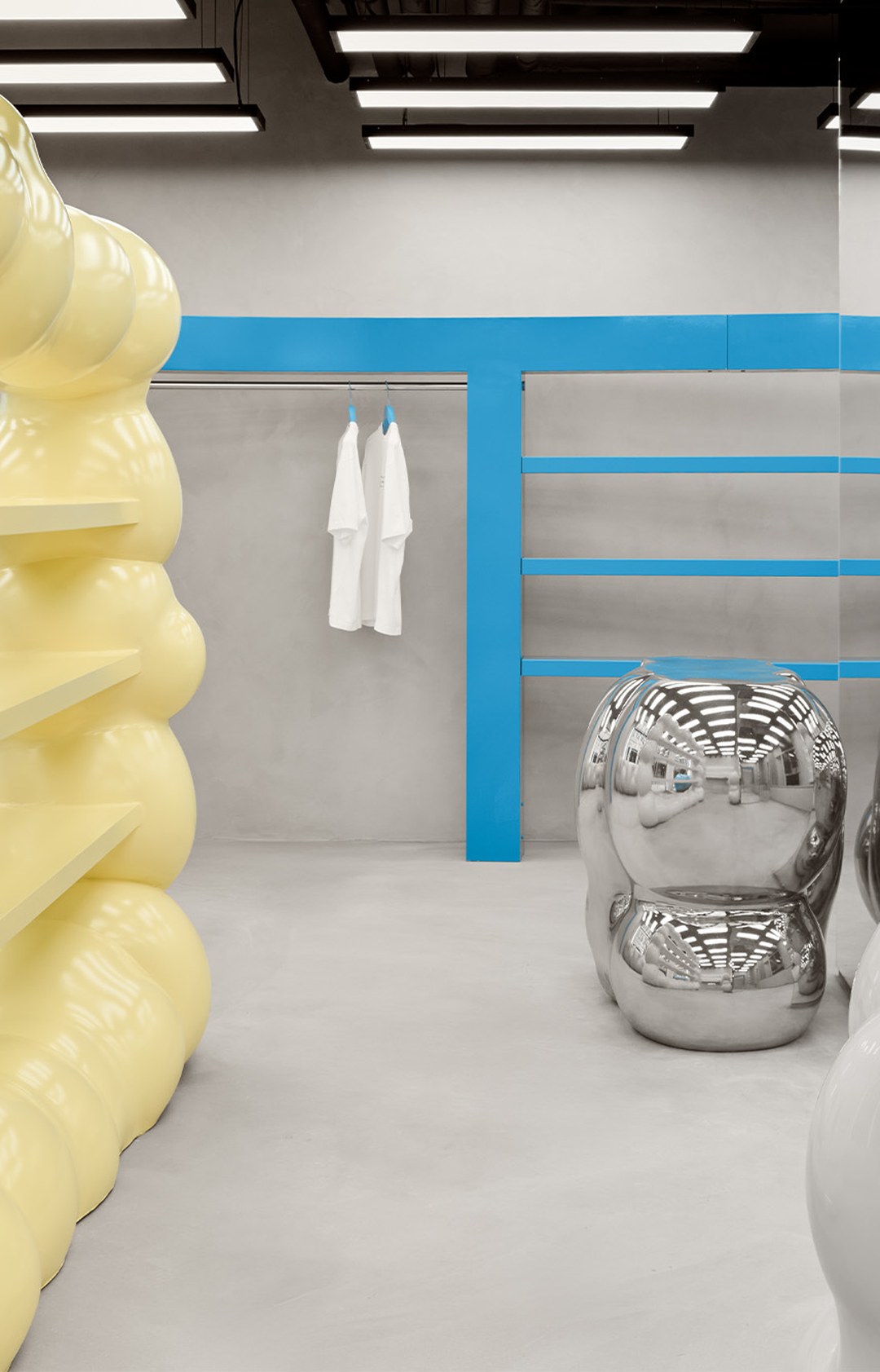 Modern retail space with yellow wavy shelves, a shiny silver stool, and minimalistic blue accents; white shirts hang in the background.