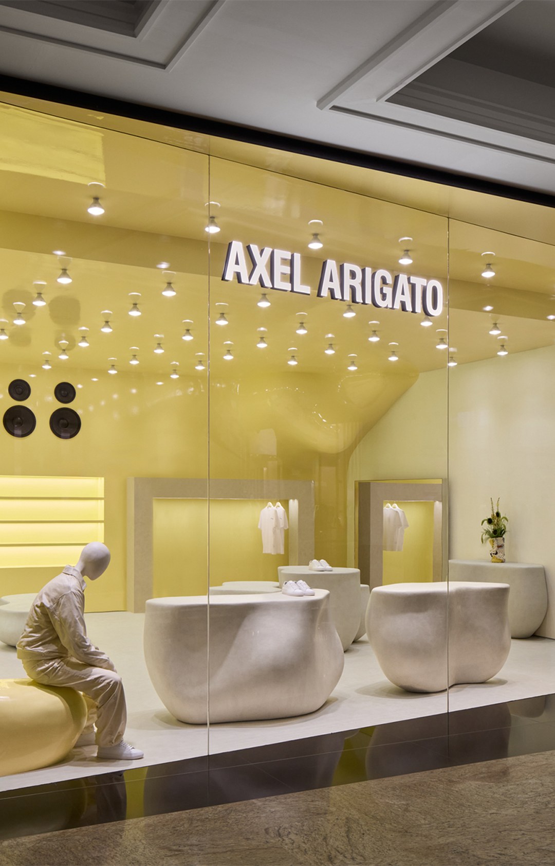 Storefront with "Axel Arigato" sign, featuring modern, minimalist interior design in yellow tones and unique stone-like furniture.