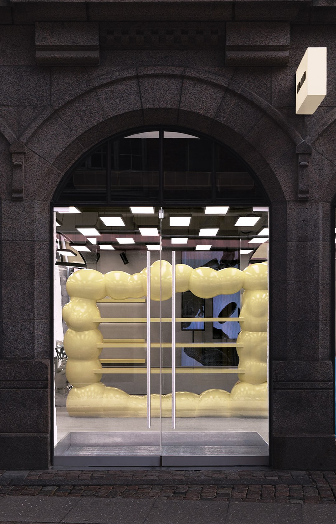 Storefront with glass doors, framed by an archway, featuring an oversized yellow balloon-style art installation inside.