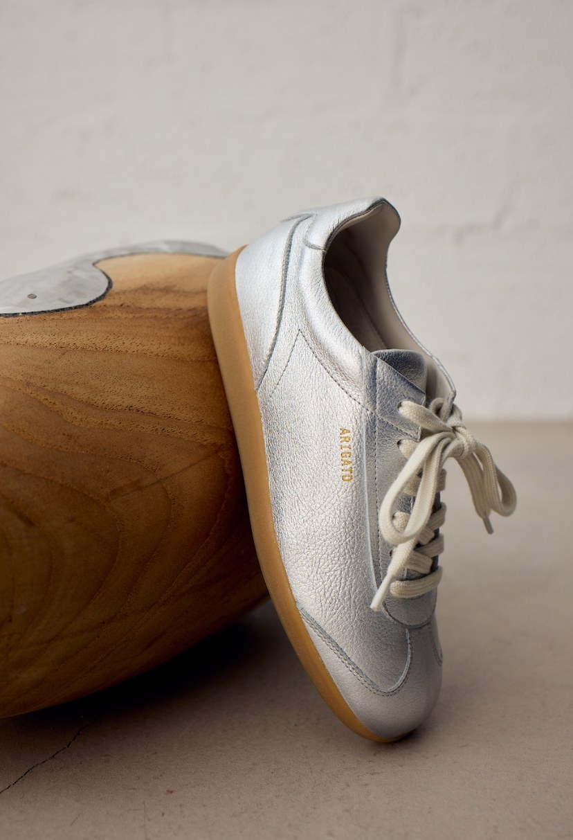 Silver leather sneaker with beige laces and gum sole, displayed against a wooden surface indoors.