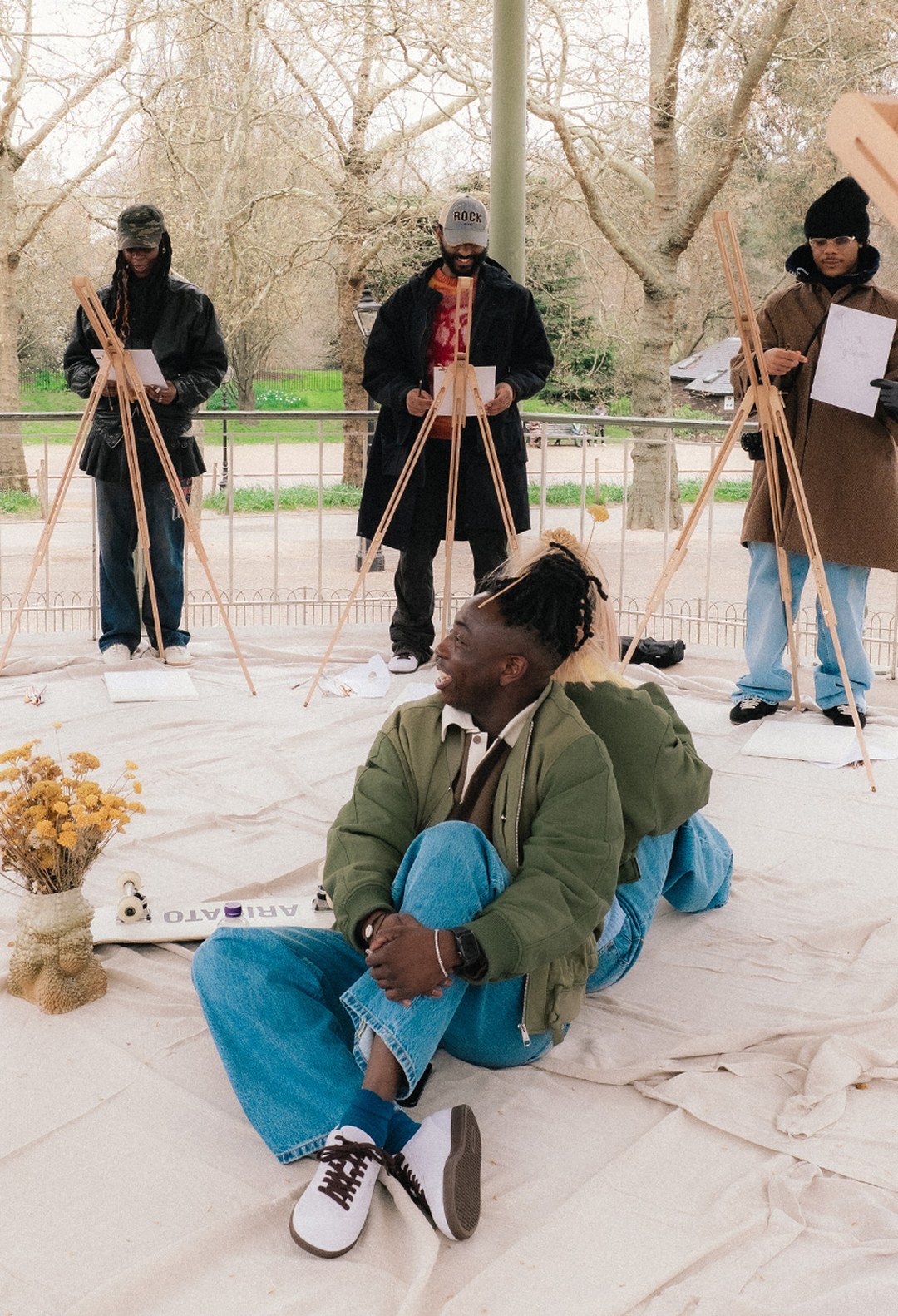 People painting on easels outdoors; one sitting on the ground, smiling, with art supplies and dried flowers nearby.