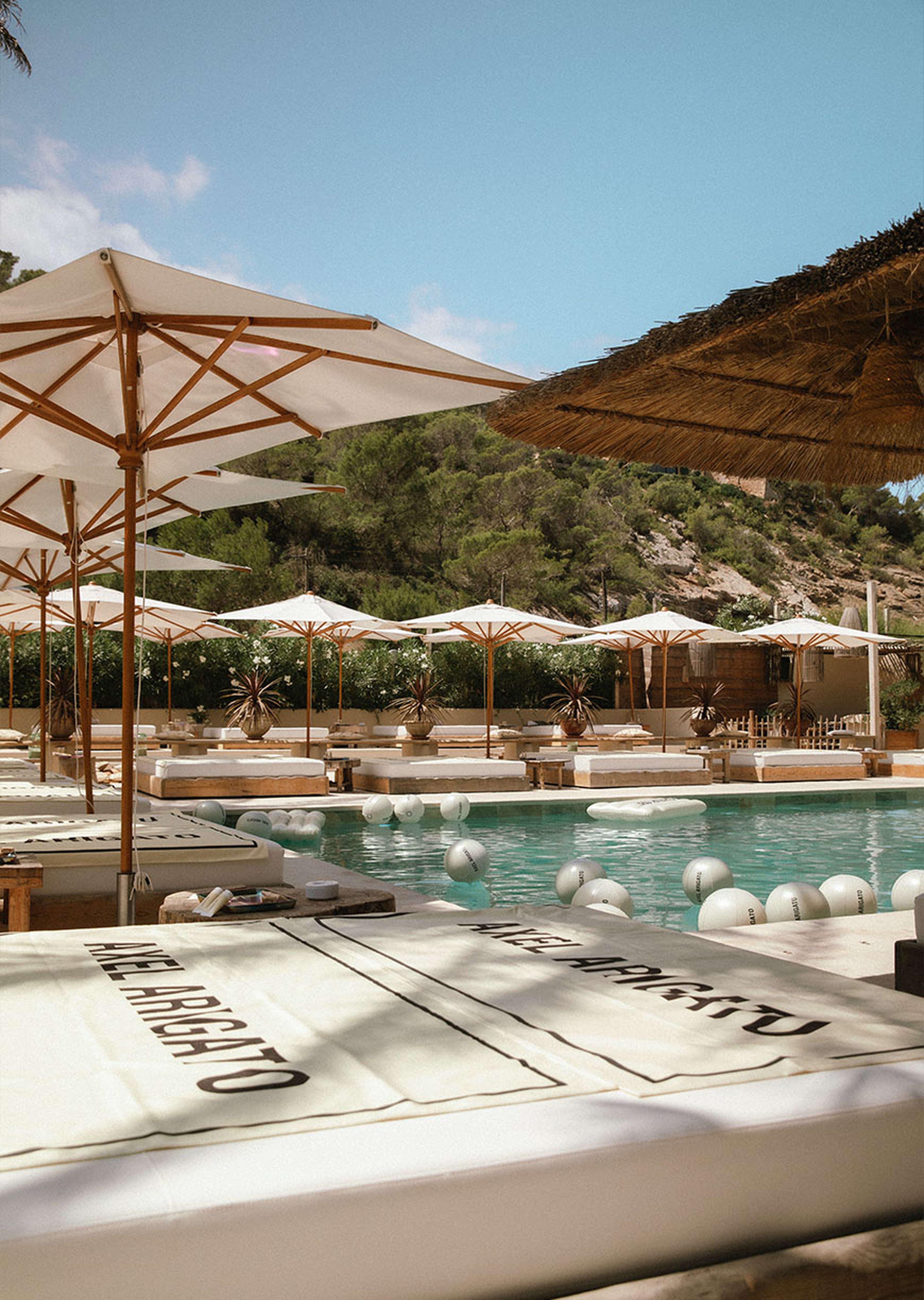 Luxurious poolside scene with white sunbeds, large umbrellas, and a clear blue pool surrounded by lush greenery under a bright sky.