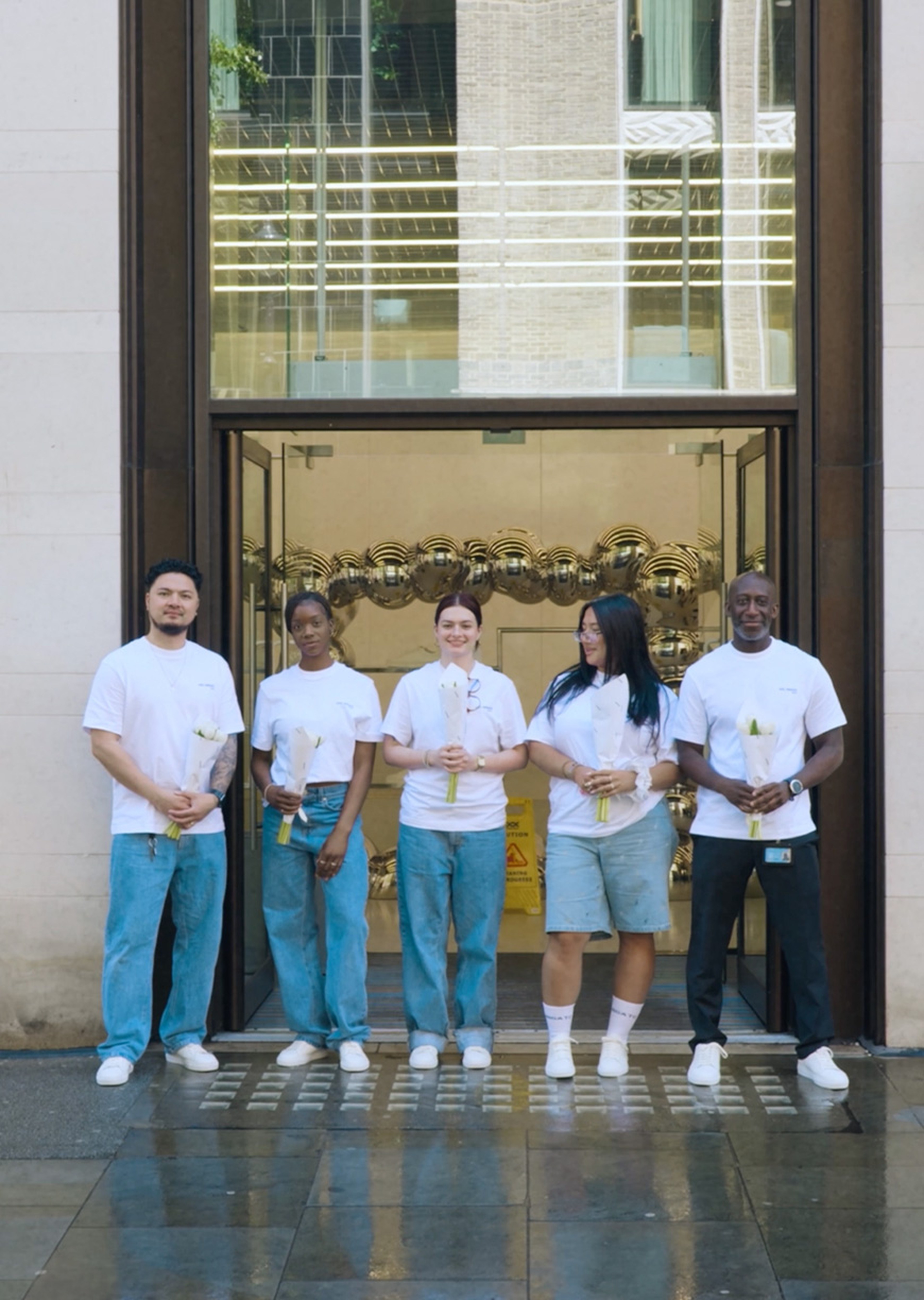 Five people in white shirts and jeans stand holding trophies in front of a glass door with gold balloons inside.