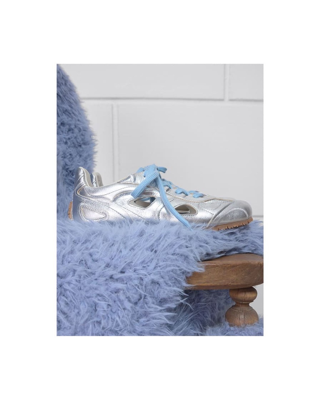 Silver sneaker with blue laces on a wooden chair draped in fluffy lavender fabric. Minimalistic white wall background.