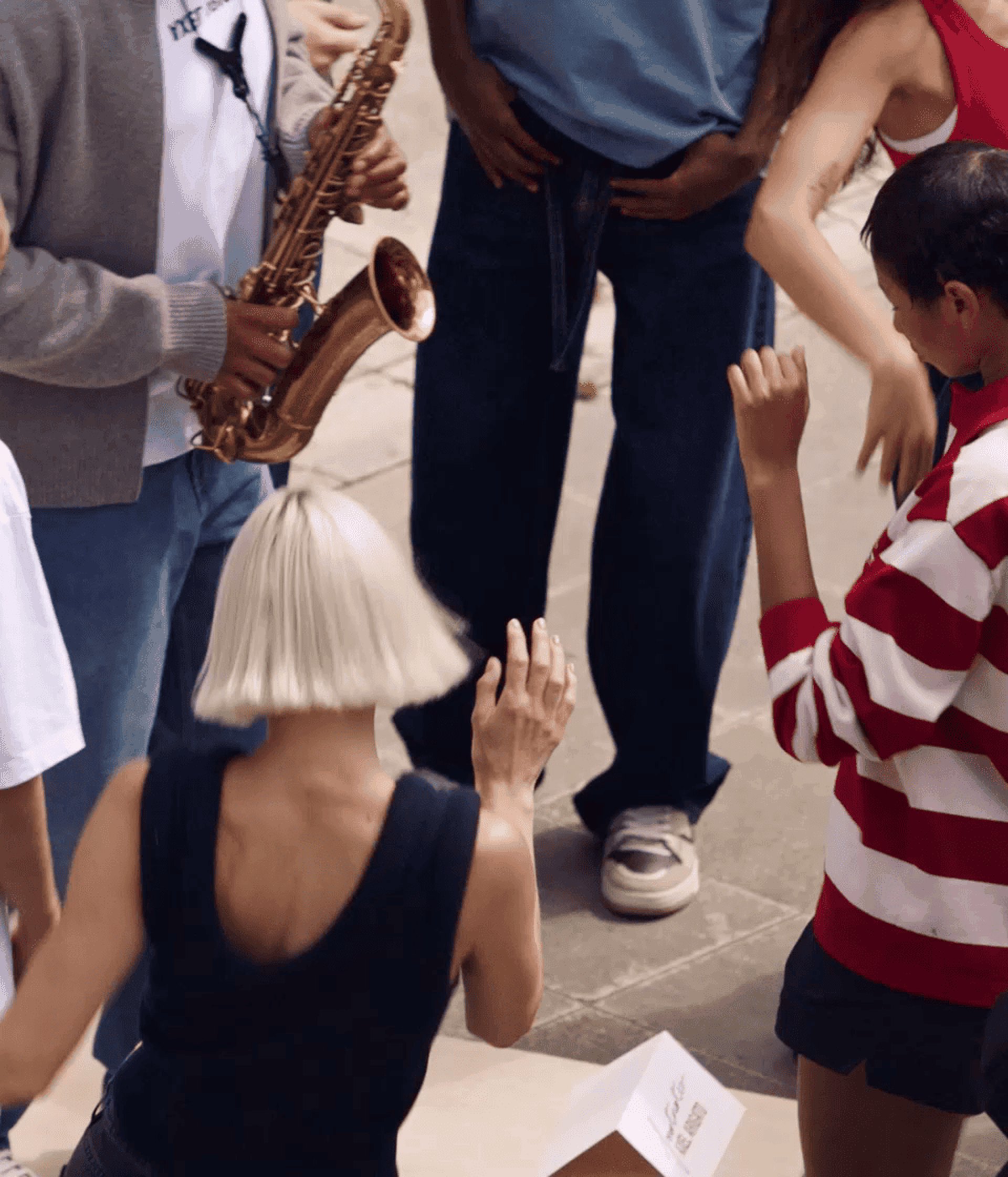 People dancing energetically outdoors, with a saxophonist playing nearby. One person wears a striped shirt, and another has short blonde hair.