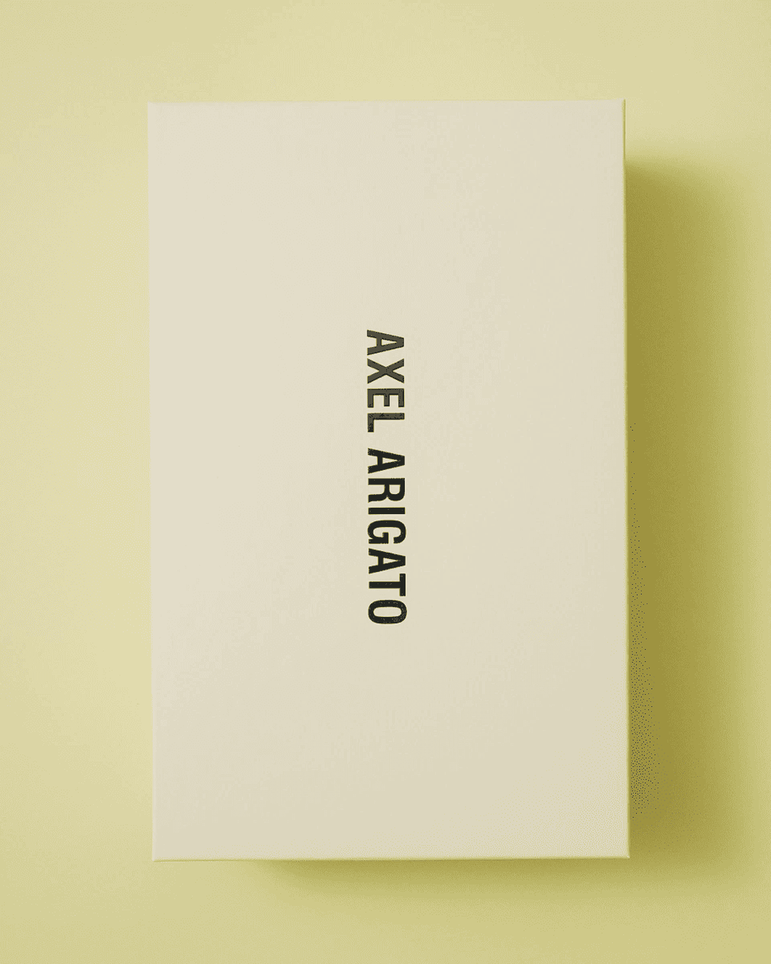 Rectangular cream-colored shoe box with "AXEL ARIGATO" text in black, lying on a light yellow background.