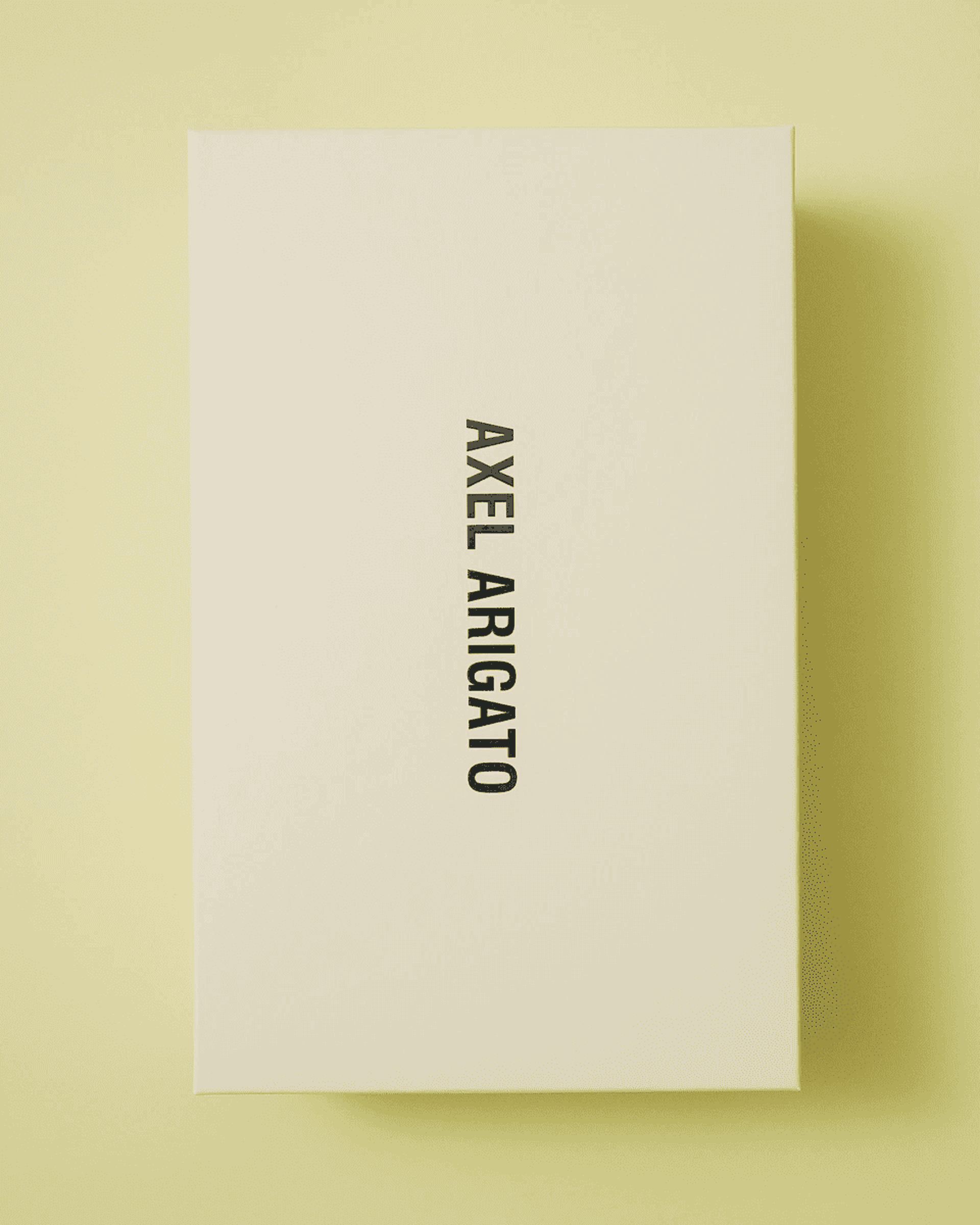 Rectangular cream-colored shoe box with "AXEL ARIGATO" text in black, lying on a light yellow background.