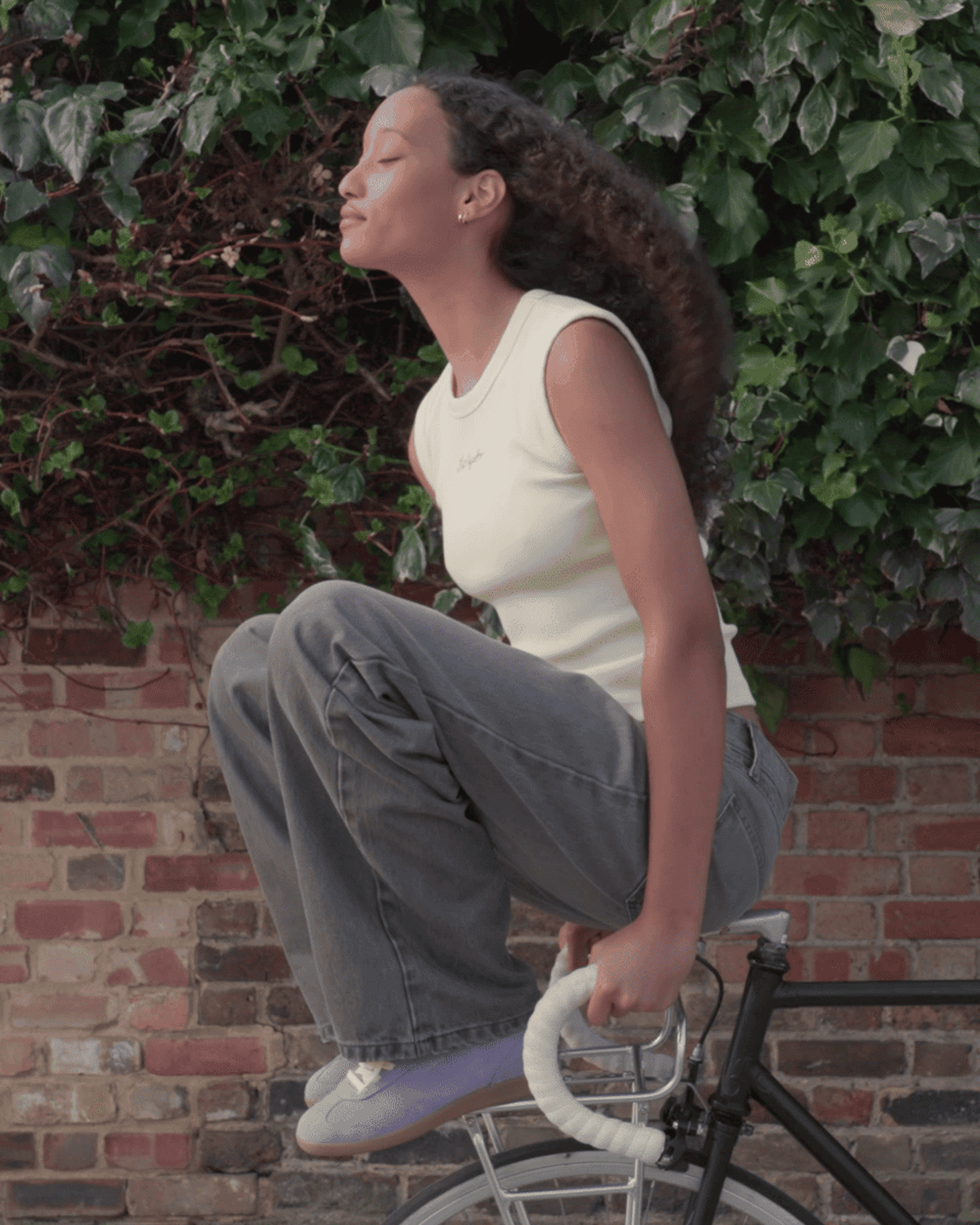 A woman in a white tank top and jeans sits on a stationary bike, eyes closed, with leafy background and brick wall.