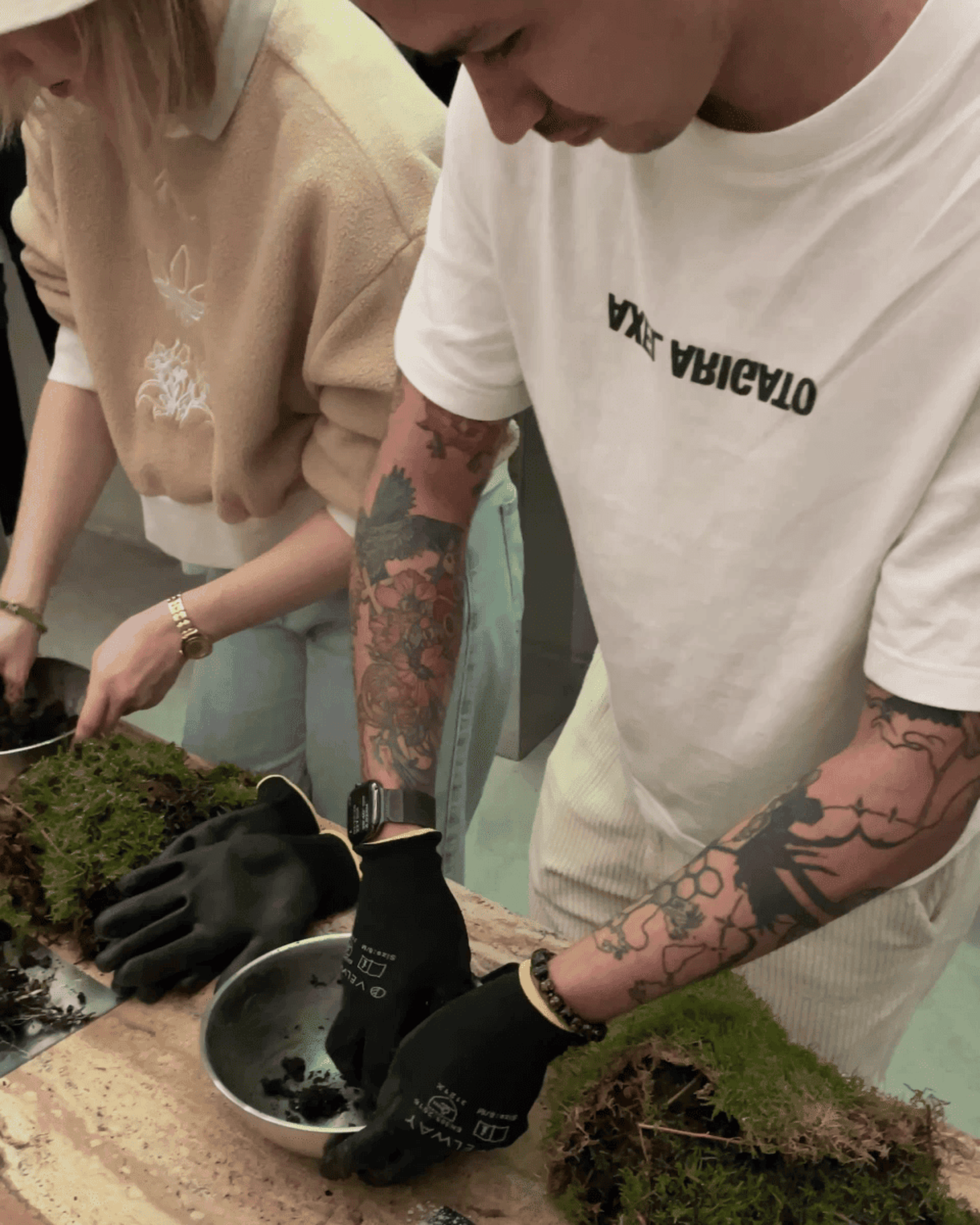 Two people arrange soil and moss on a table. The man has tattoos and wears black gloves, while the woman wears a beige sweater and gold watch.