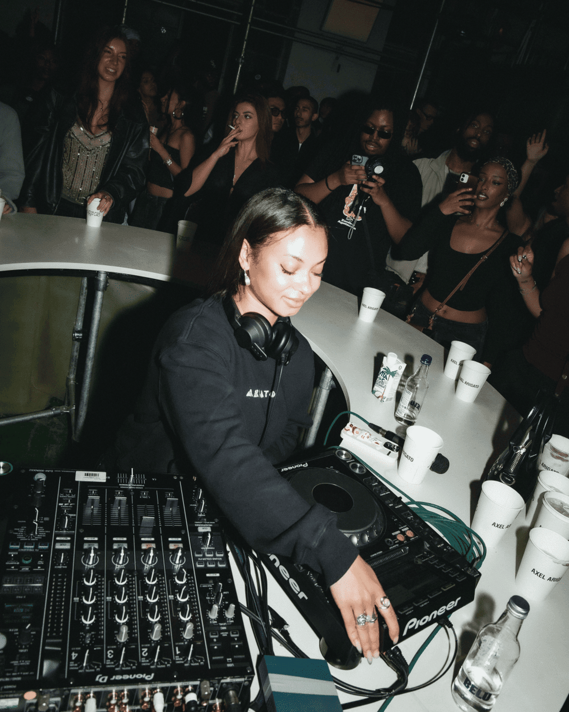DJ performing at a lively event, surrounded by people enjoying music, with audio equipment and drinks on a white table.