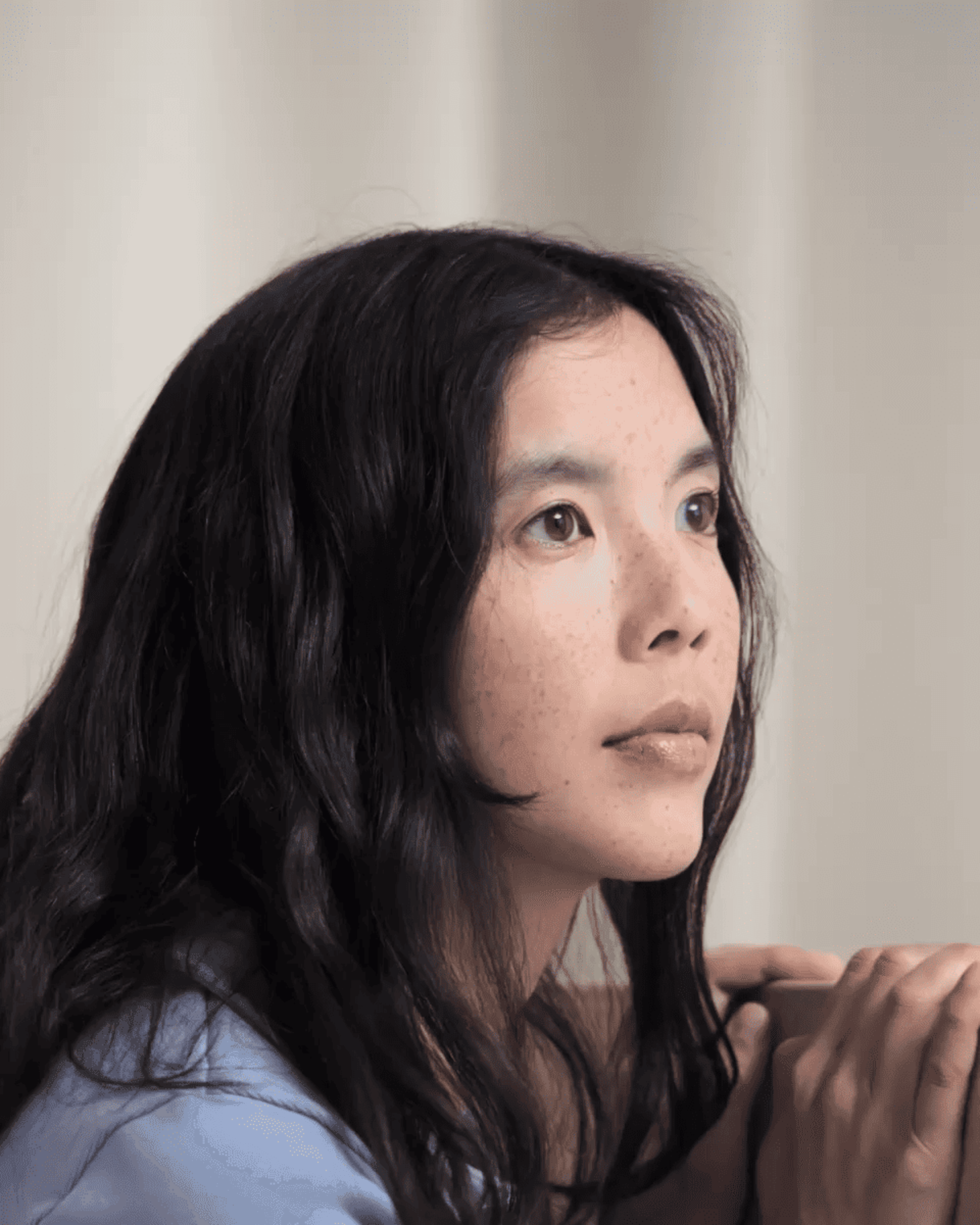 Woman with long dark hair gazes upward thoughtfully, hands clasped. Neutral background.