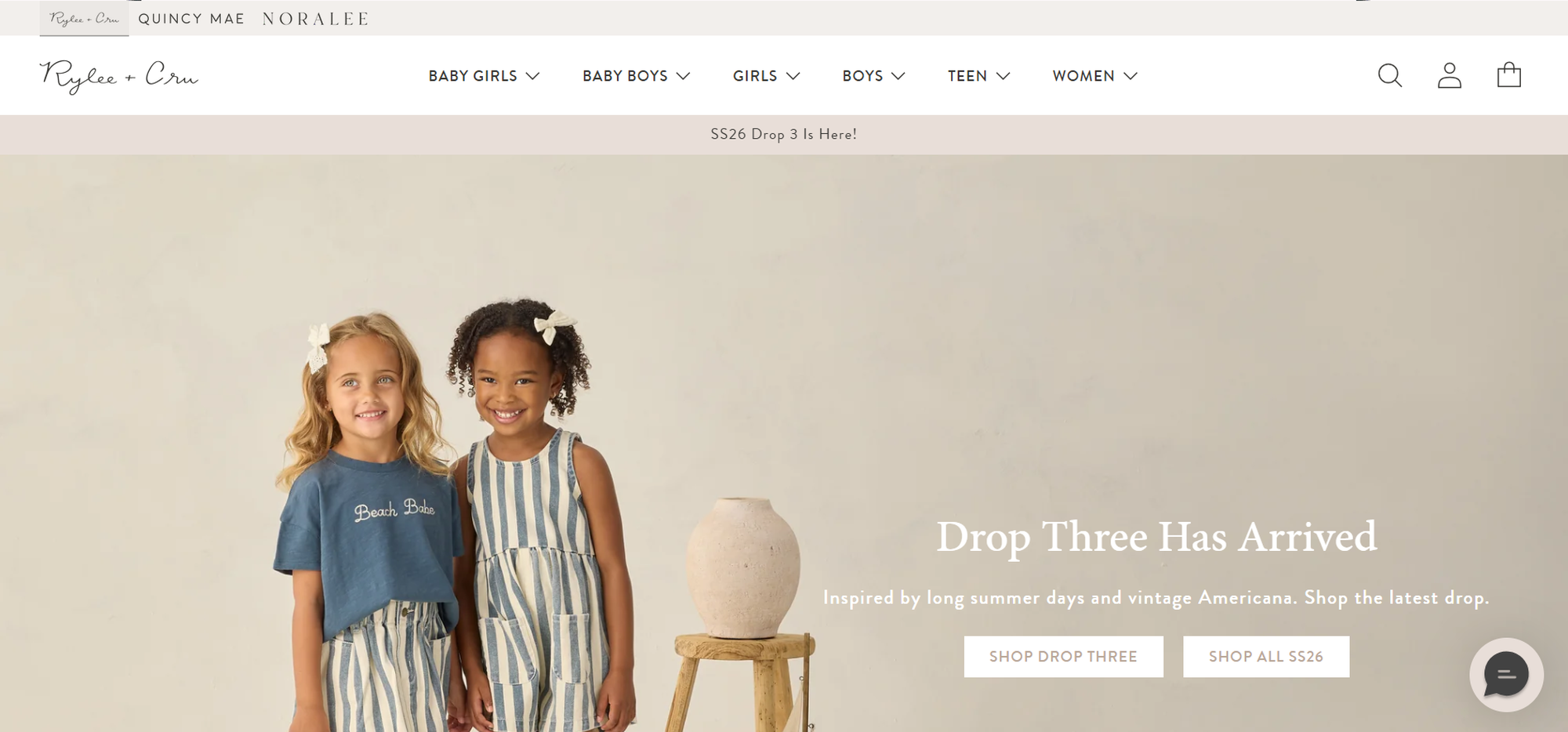 Two young girls smiling, wearing striped dresses. One has blonde hair and the other has curly black hair. They stand next to a wooden stool with a vase.