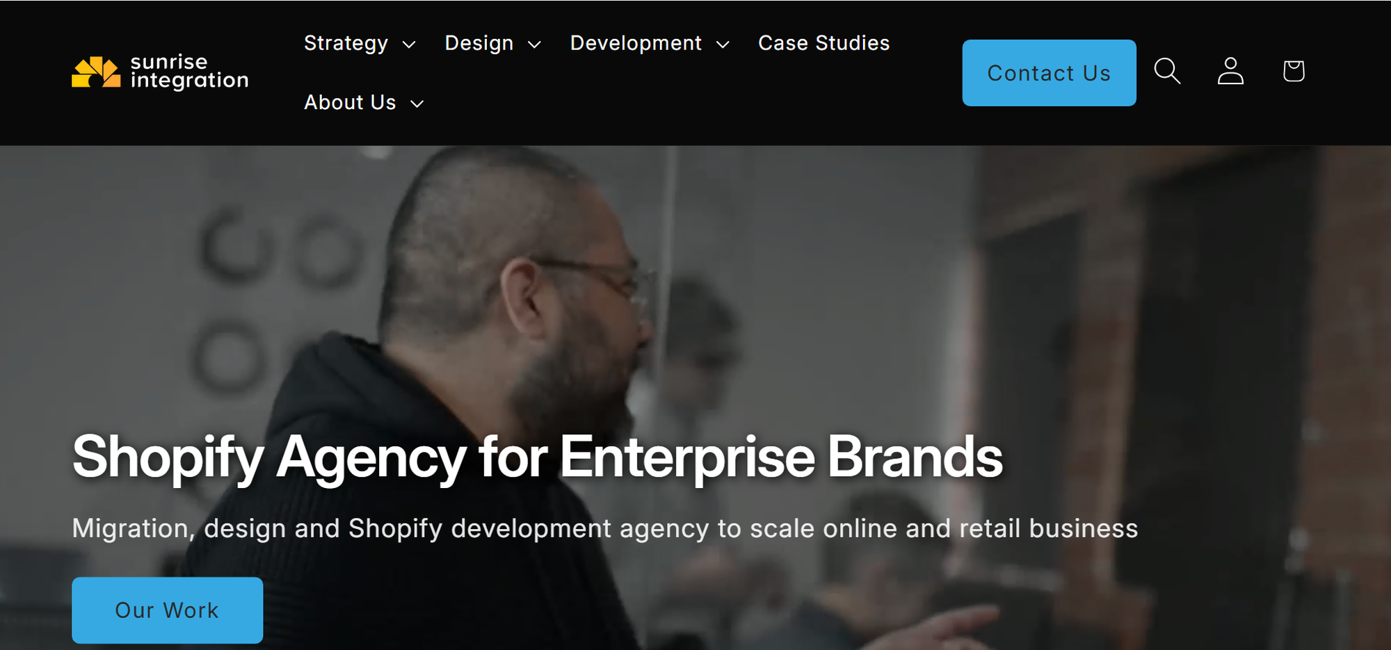 Man gesturing while speaking in a professional setting. Text overlay: "Shopify Agency for Enterprise Brands" with navigation menu and contact button.