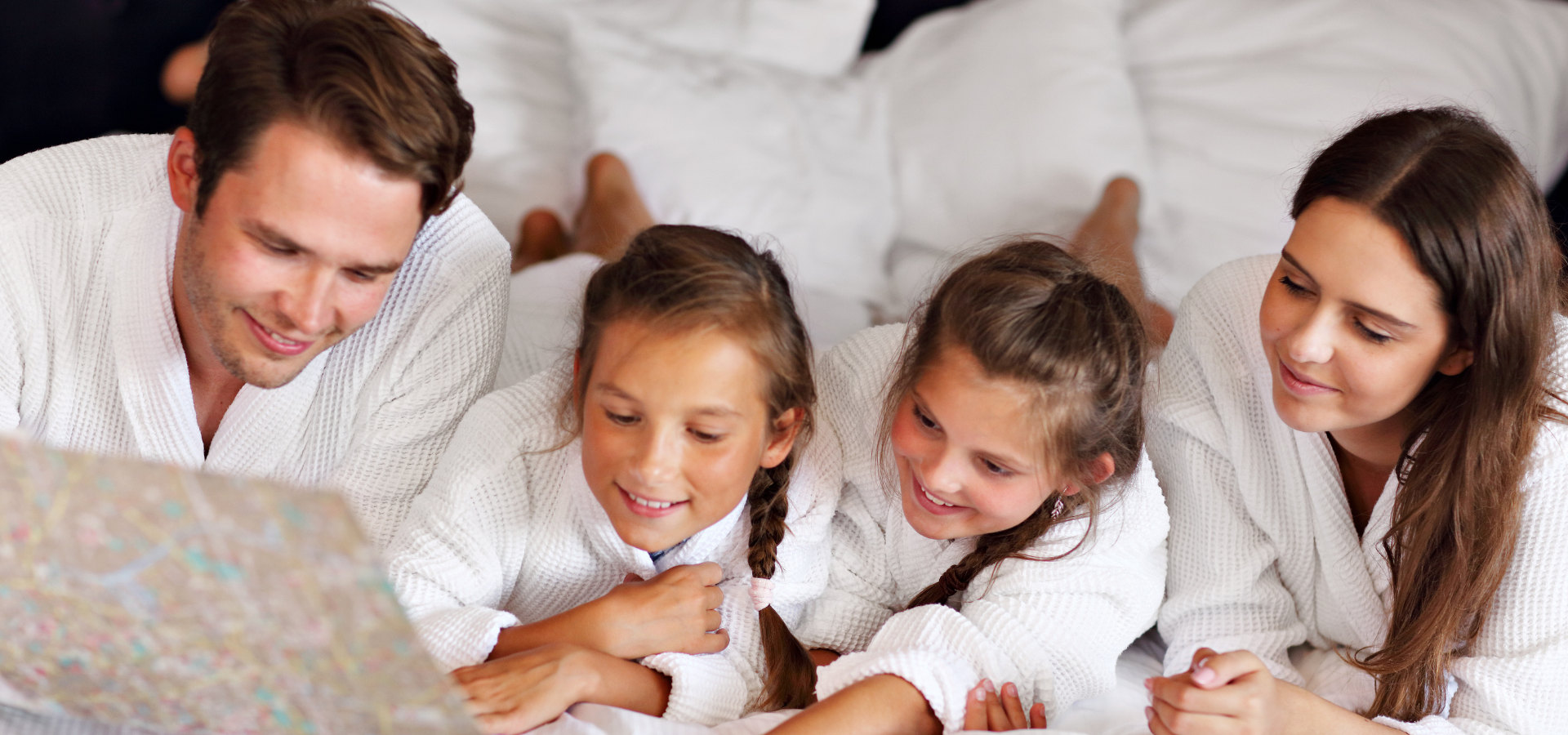 Family of four in white robes lying on a bed, smiling as they look at a map or book together.