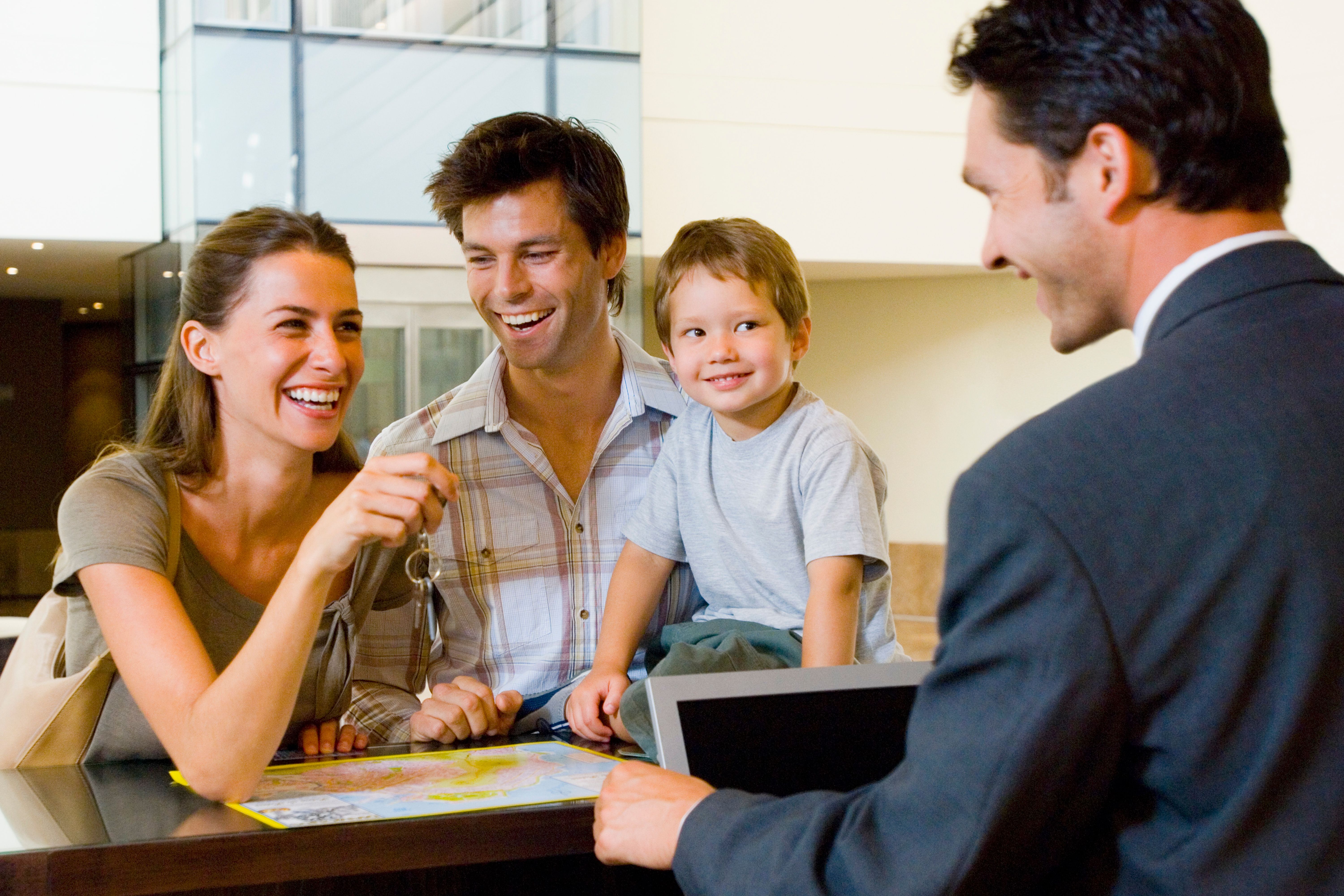 Smiling young family (mother, father, son) receiving keys and paperwork from a smiling agent at a desk.