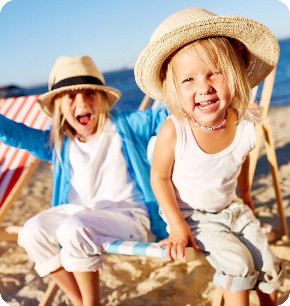 Two smiling children in sun hats enjoy a sunny beach day, sitting on deck chairs with the ocean and a striped beach chair in the background.