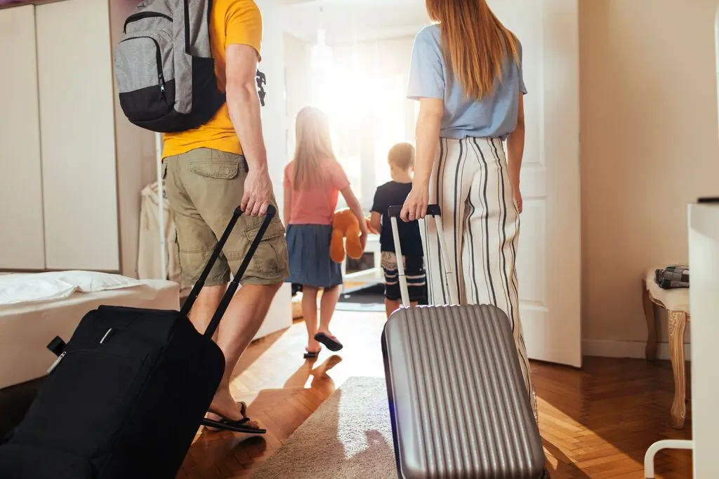 A family of four, seen from behind, carrying luggage as they walk through a sunlit room, heading towards a door, suggesting travel or departure.