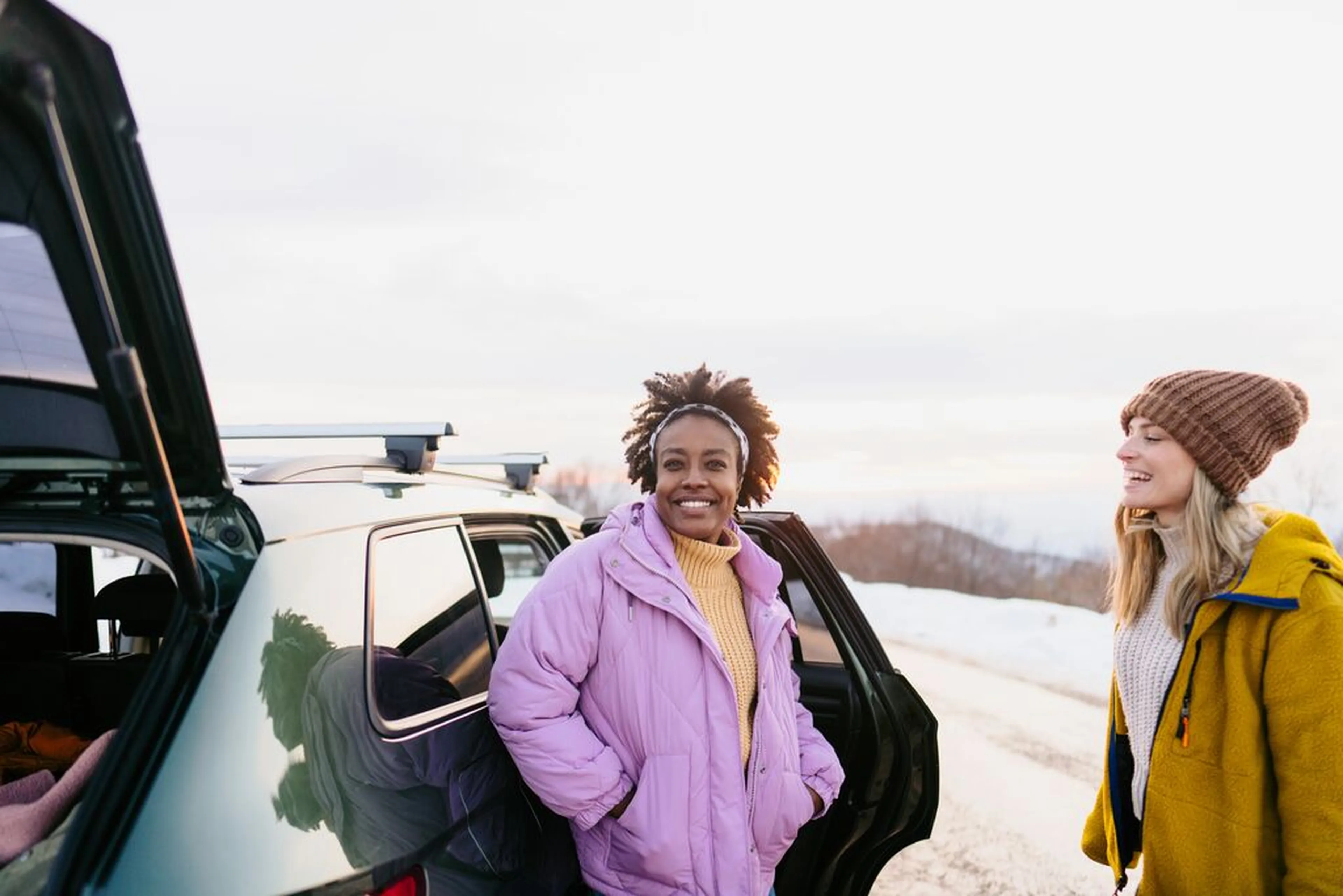 Two women in winter coats stand smiling by an open car door on a snowy roadside with a scenic background.