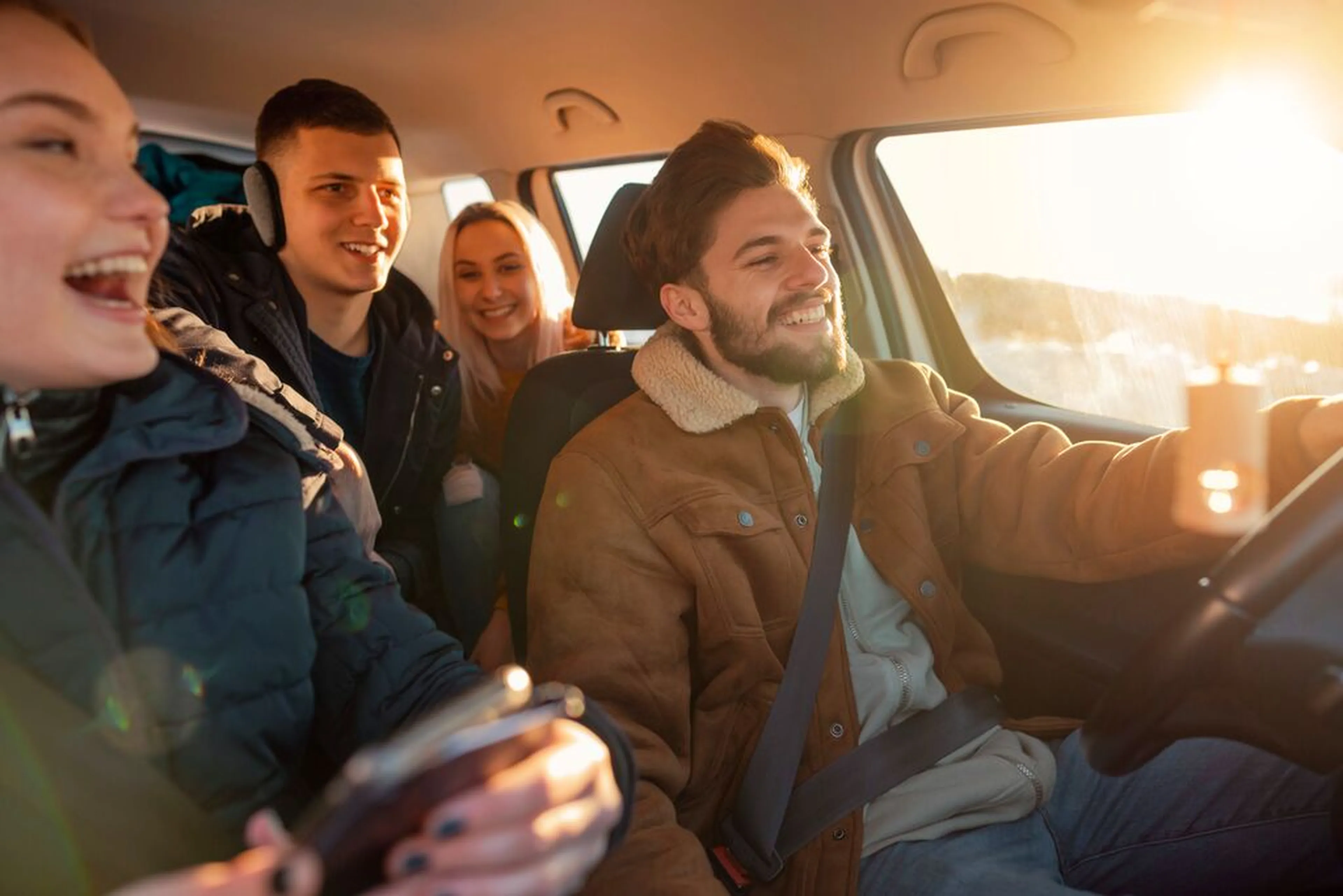 Four friends wearing coats are joyfully driving in a car, with the sun setting outside.