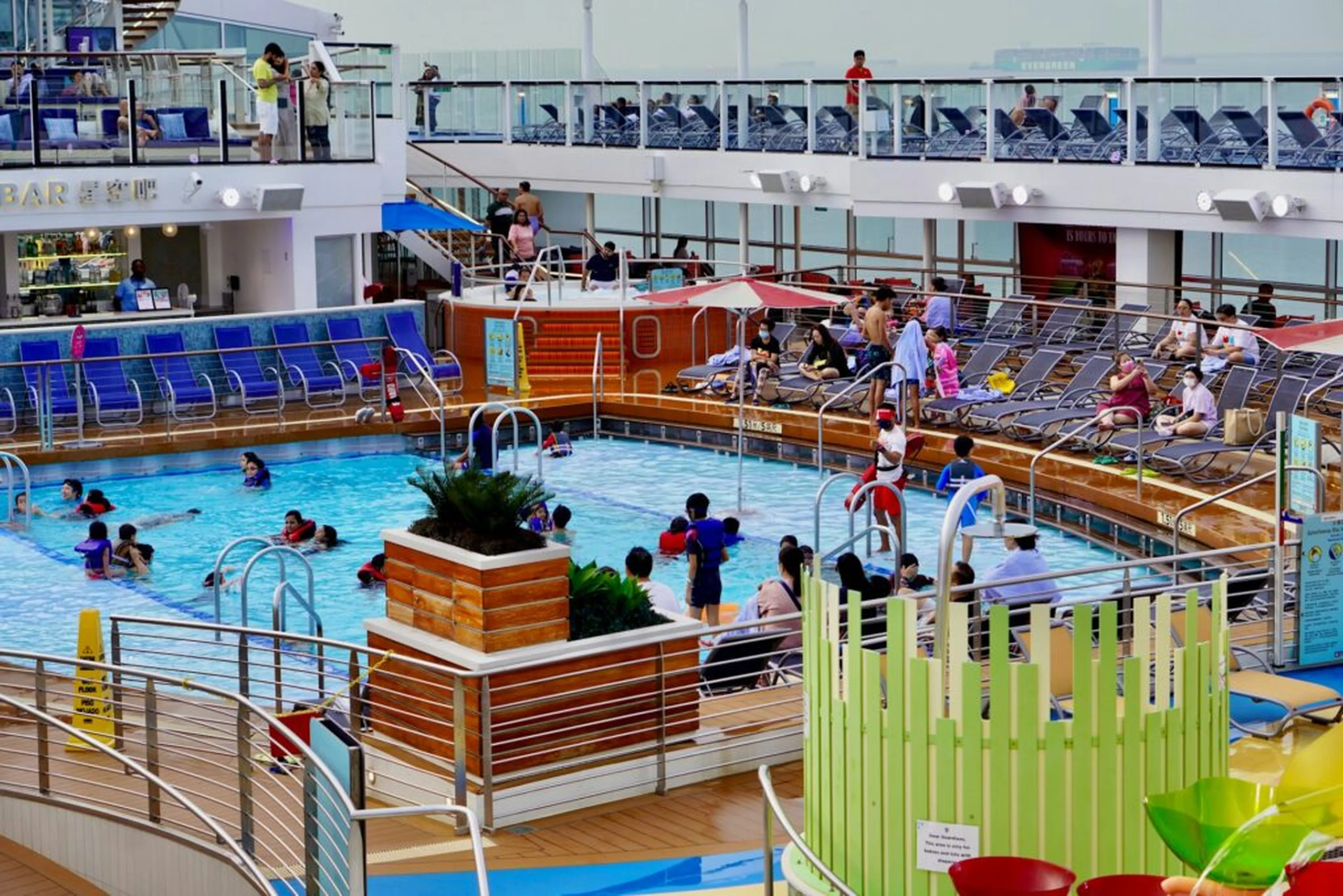 People enjoying a swimming pool on a cruise ship deck, surrounded by loungers and sunbathers, with a bar and colorful decor nearby.