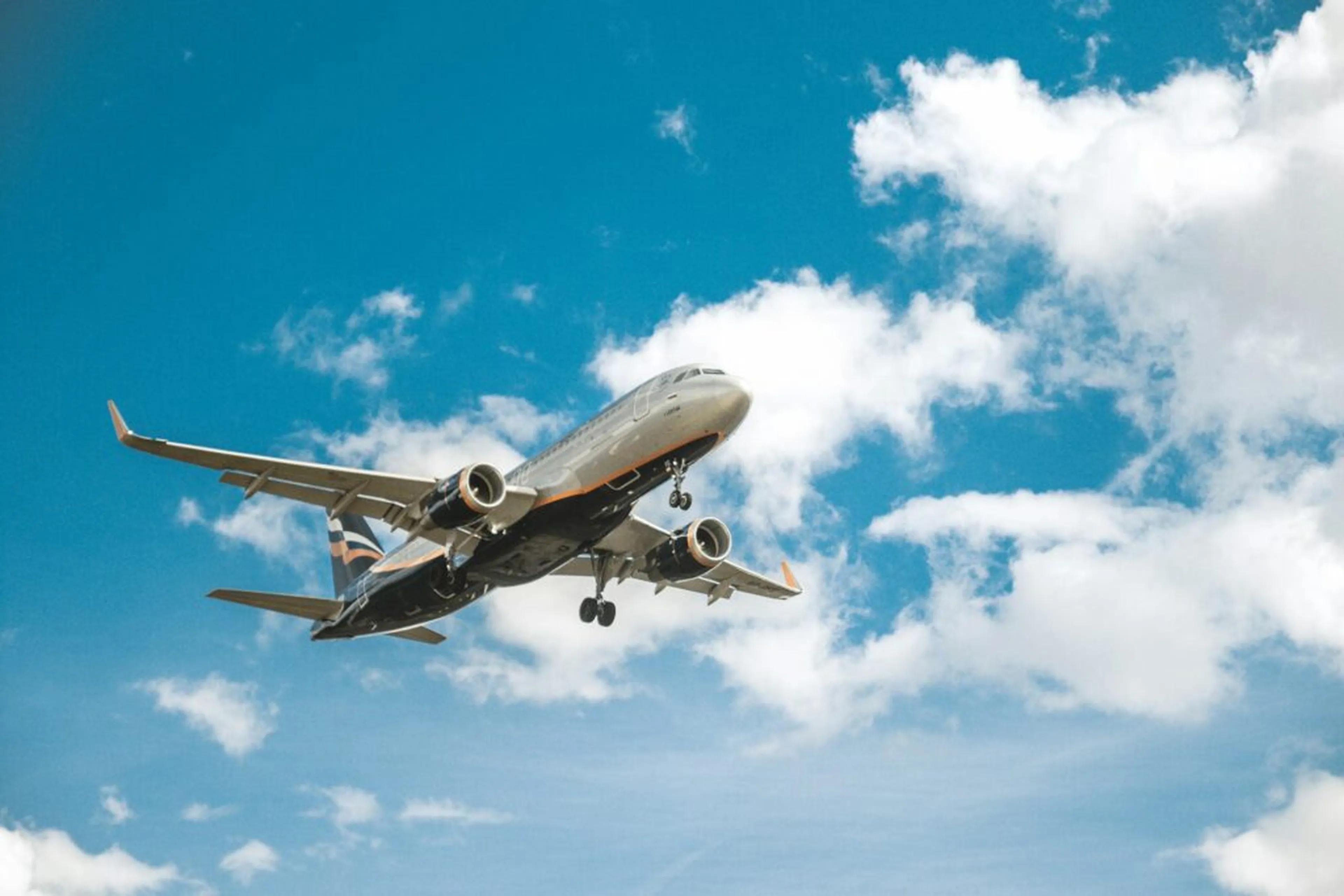 An airplane flies low in a clear blue sky, surrounded by fluffy white clouds.