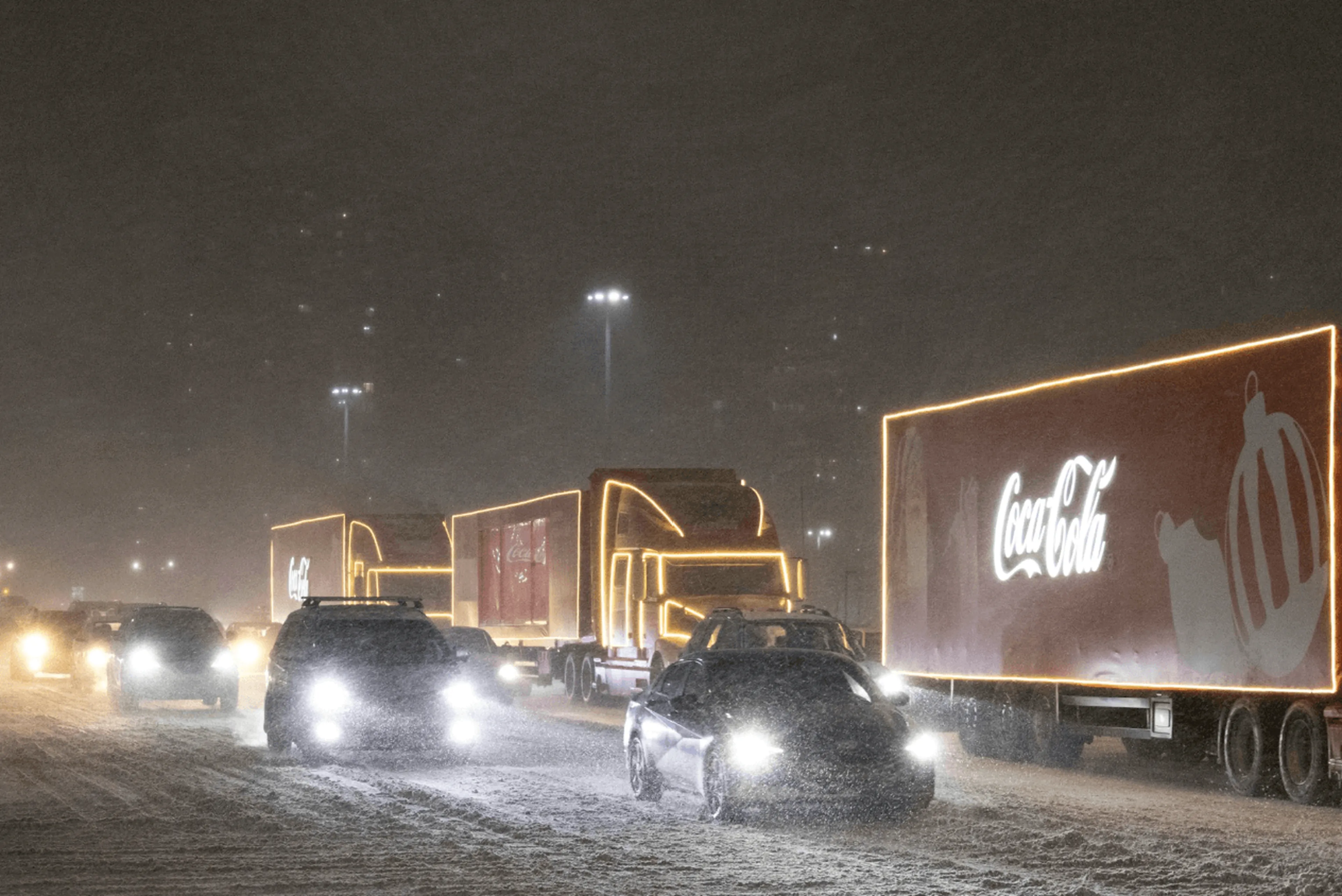 Trucks and cars drive through heavy snow at night, with two illuminated Coca-Cola trucks visible in the traffic.