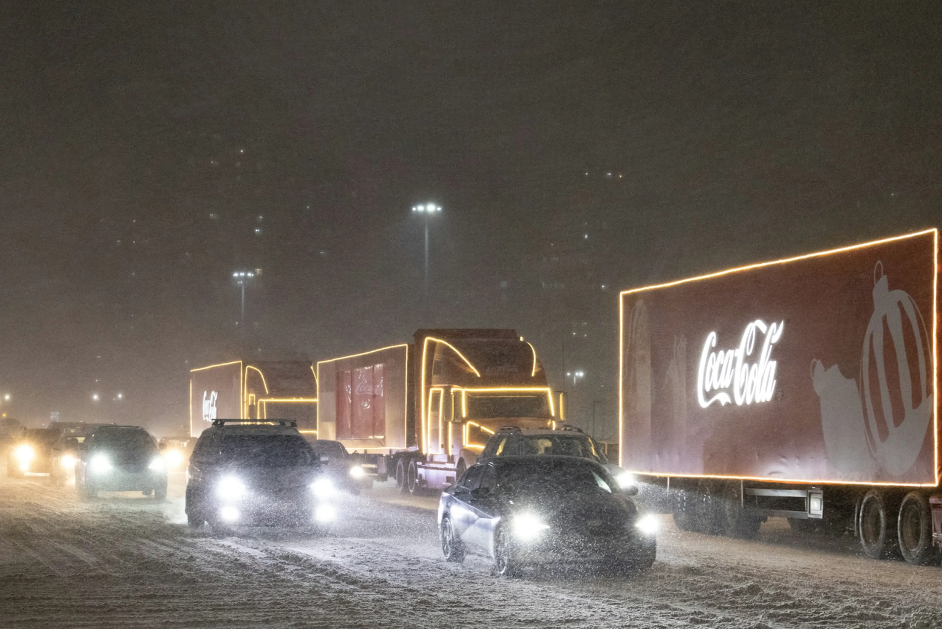 Coca-Cola trucks and cars drive through a snowy street at night, with bright lights illuminating the scene.