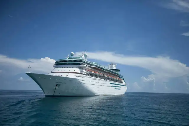 A large white cruise ship sails on a calm ocean under a clear blue sky with scattered clouds.
