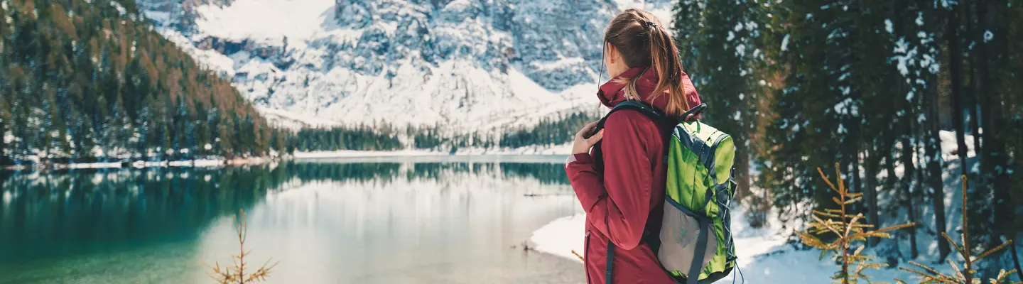 Hiker in a red jacket with a green backpack gazes across a turquoise alpine lake toward snow-covered mountains and a pine forest.