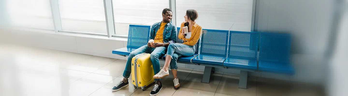 A couple sits on blue chairs in an airport, smiling and chatting. A yellow suitcase is nearby, and the woman holds a coffee cup.