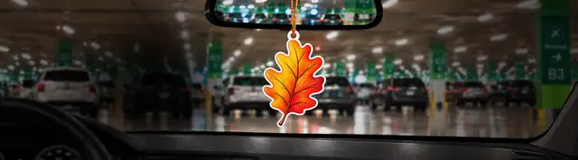 A car's interior with an orange leaf-shaped air freshener hanging on the rearview mirror, overlooking a busy parking garage.