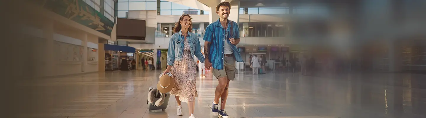 A couple casually strolls through an airport terminal, smiling and holding hands, with a hat and a wheeled suitcase in tow.