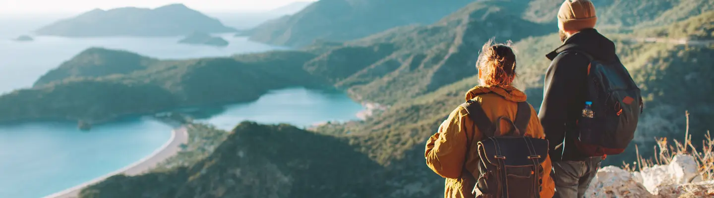 Two hikers with backpacks on a ridge overlooking a curved beach, turquoise bay and forested islands beneath hazy mountains.